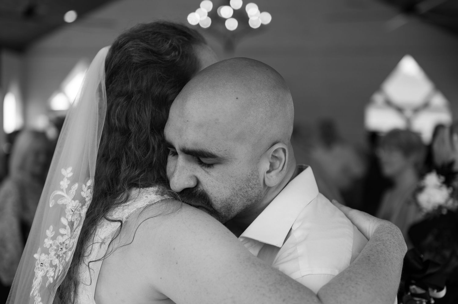 Black and white wedding photo of a bald groom embracing his veiled bride in an emotional hug.