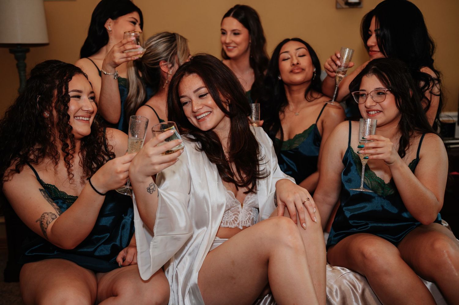 Bride in white robe takes selfie with bridesmaids in teal dresses holding champagne glasses during wedding preparations.
