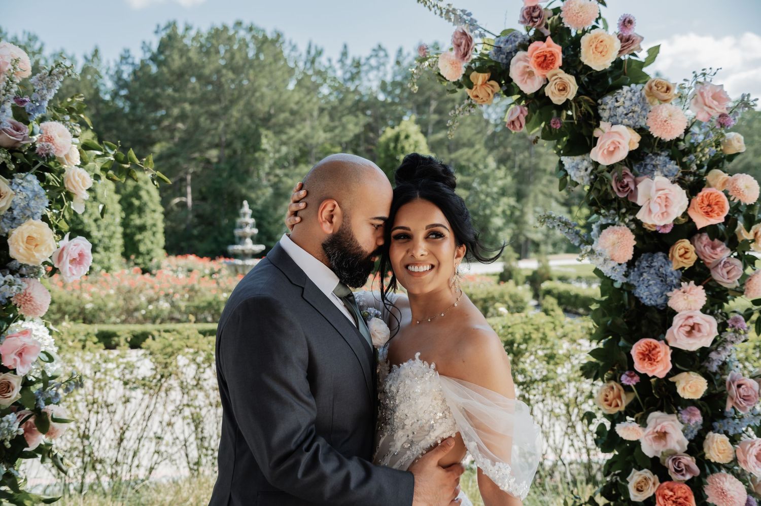 Bride and groom share a romantic moment under a lush floral arch with pink and blue blooms outdoors.