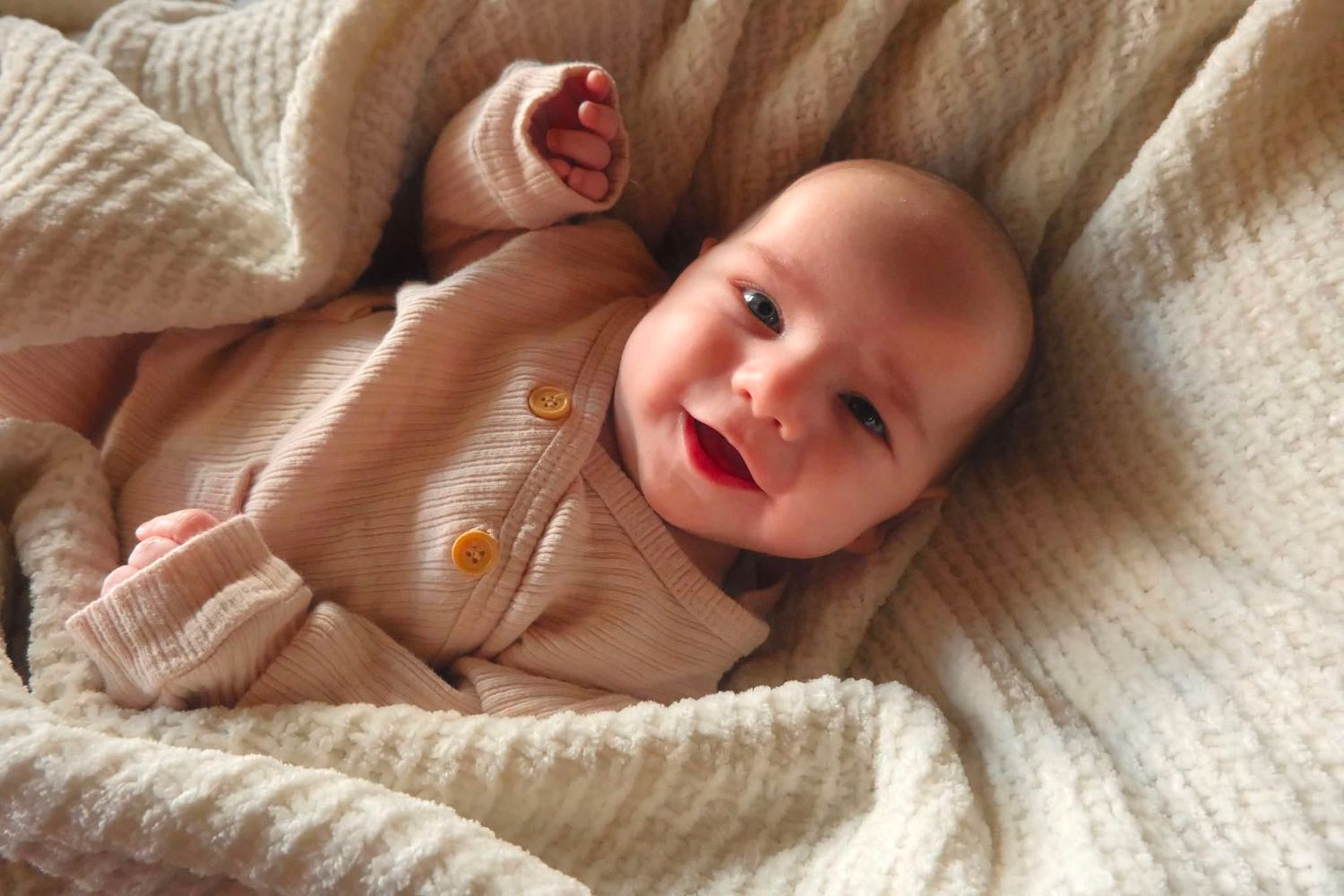 Smiling baby in beige knit outfit lying on a soft cream blanket, looking happy and content.