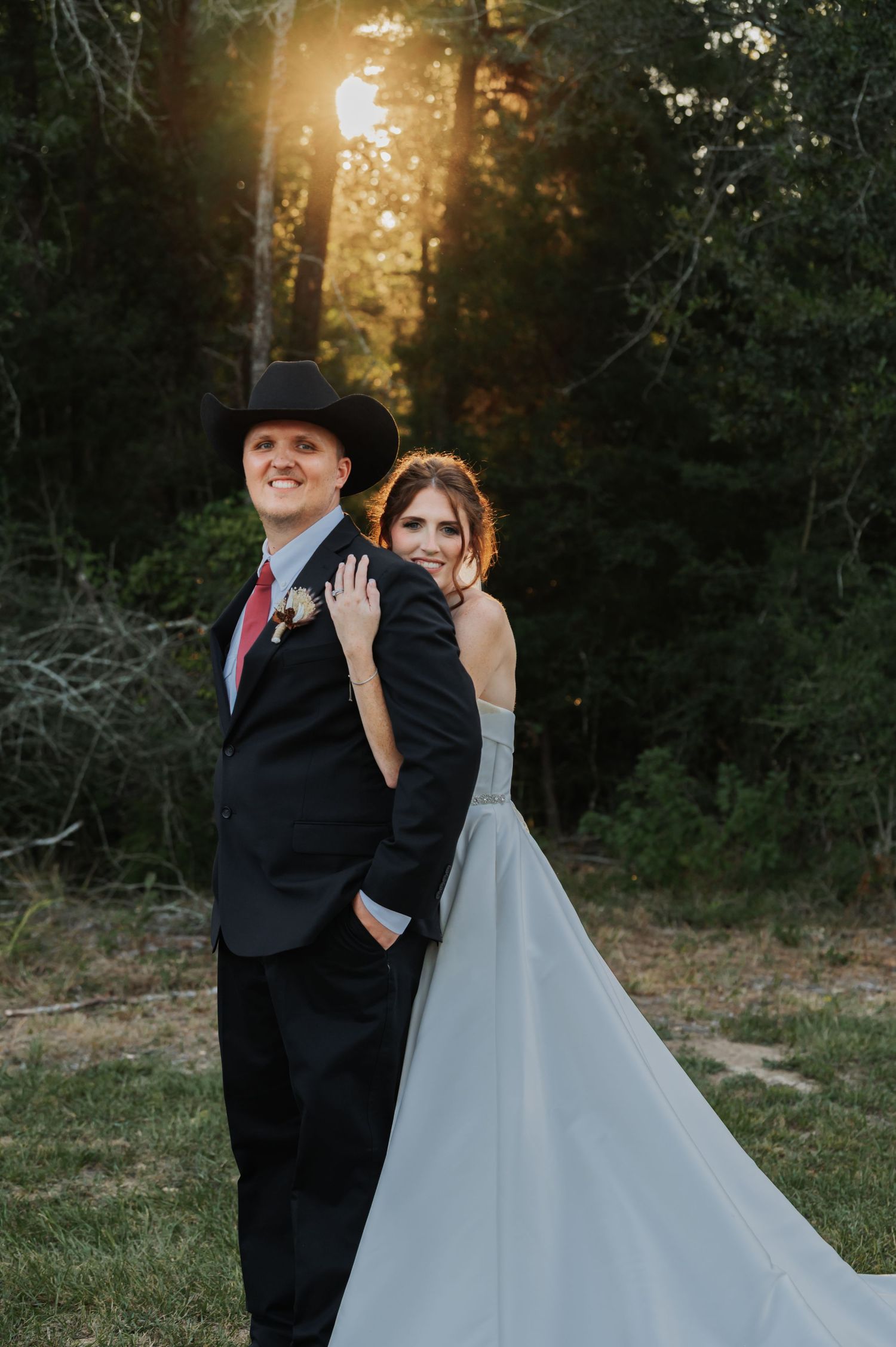 Bride embraces groom from behind as they pose together at sunset in a wooded outdoor wedding setting.