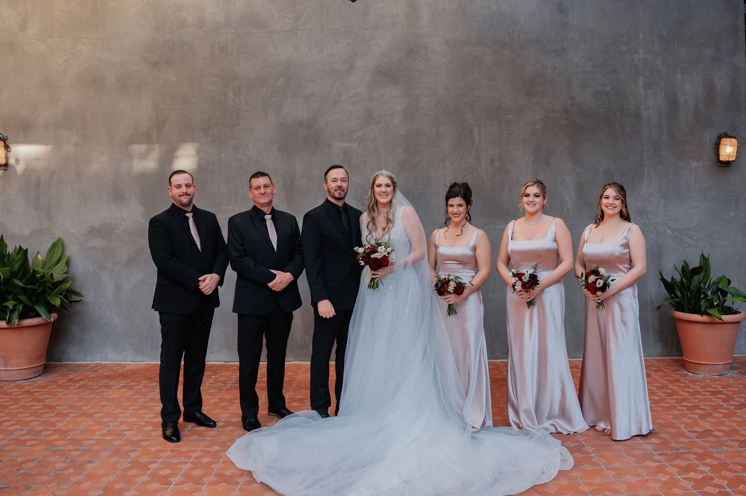 Wedding party posing together, bride in white gown with bridesmaids in blush dresses and groomsmen in black suits.