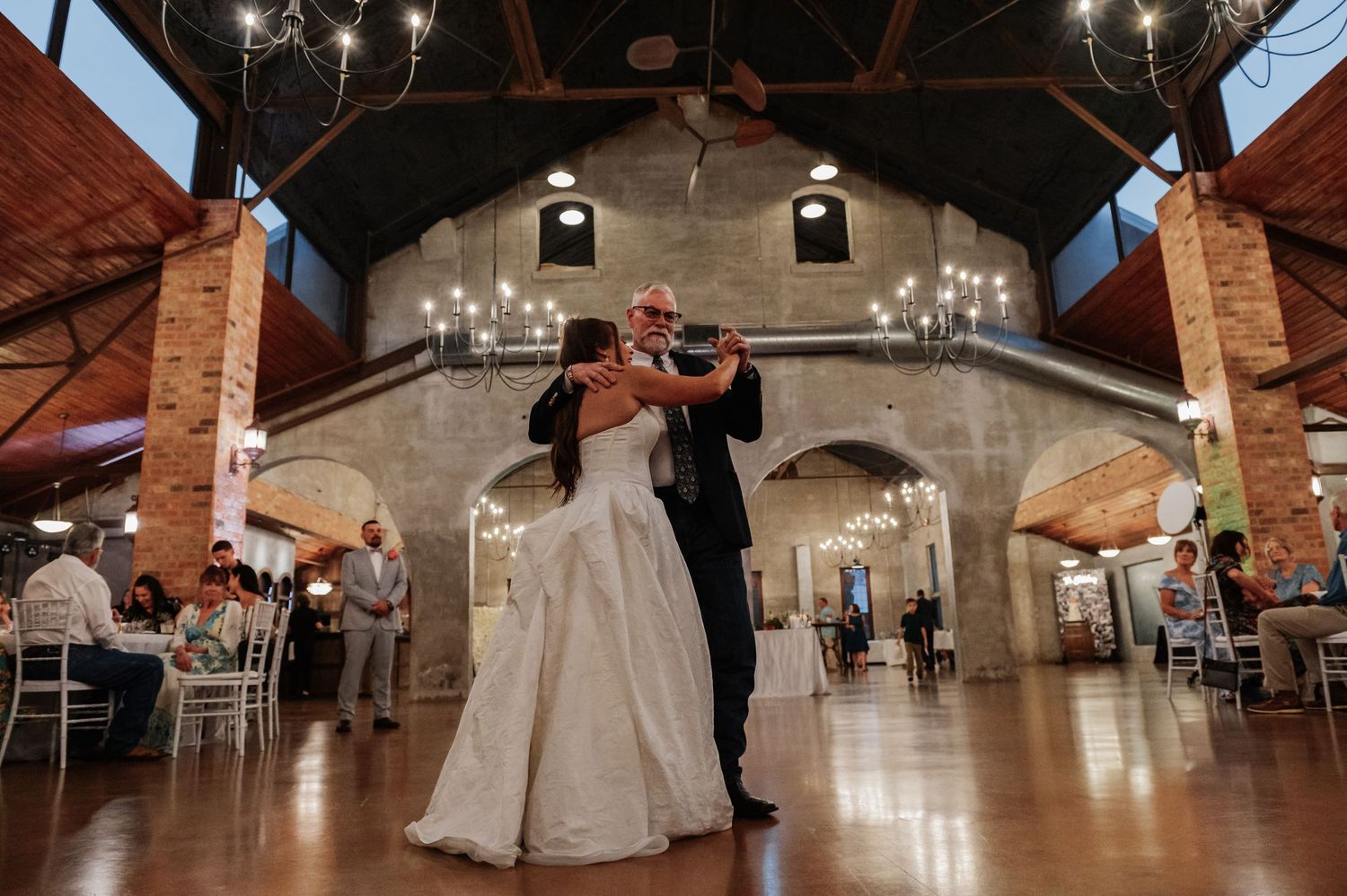 Bride and groom share first dance in elegant rustic wedding venue with arched ceilings and chandelier lighting.