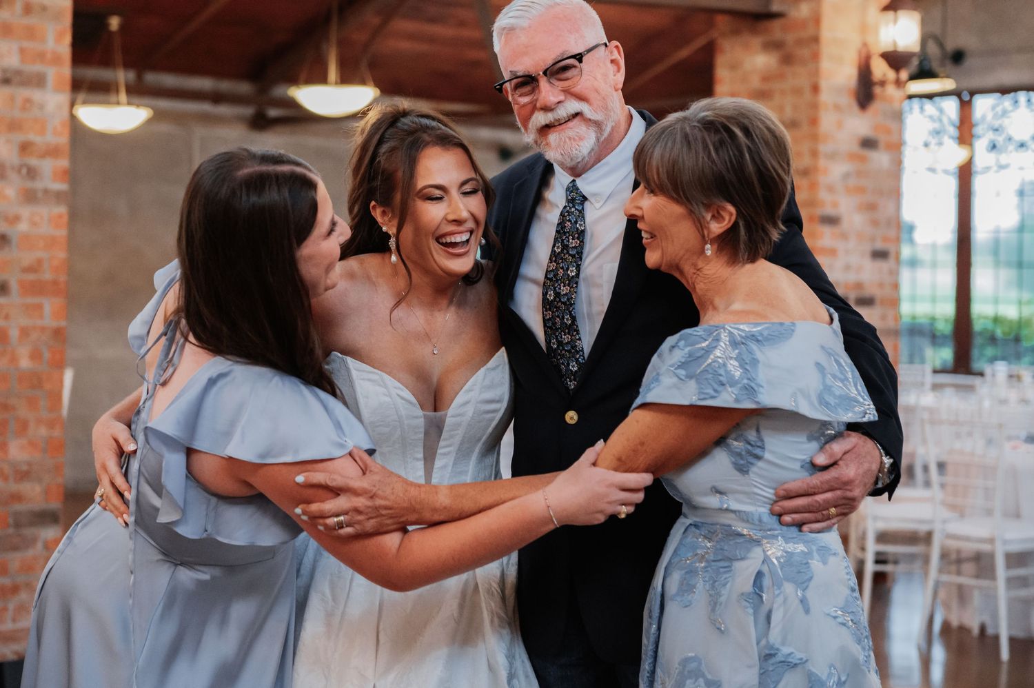 Bride in white gown hugging family members in blue outfits with older gentleman at wedding reception.