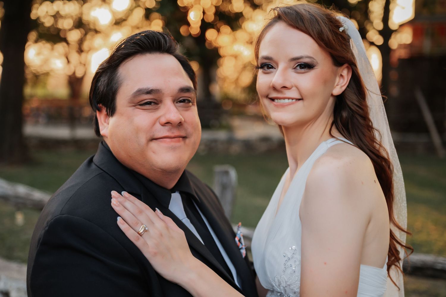 A smiling bride in white gown poses with groom in black suit at outdoor wedding reception.