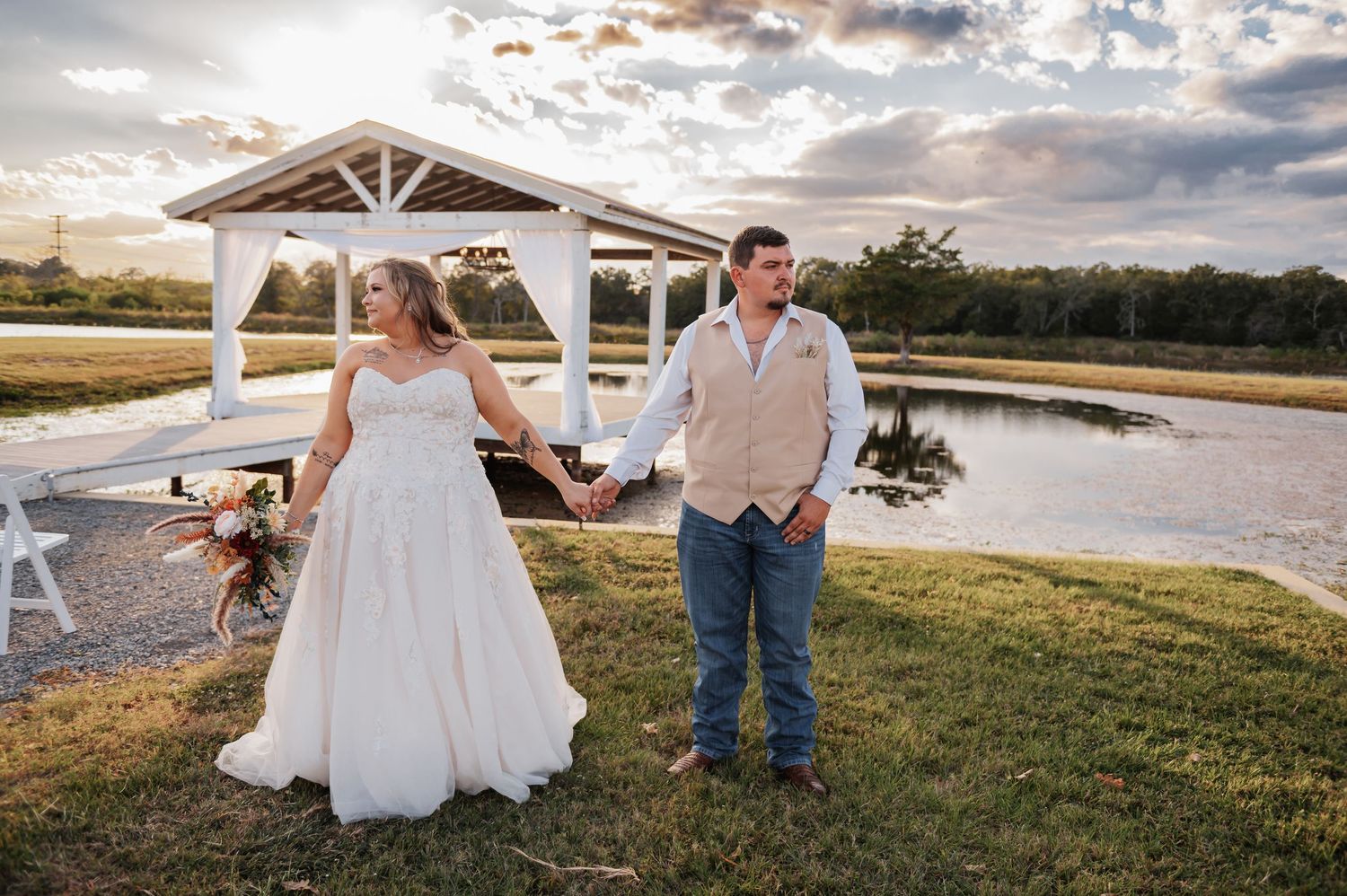 A bride and groom hold hands near a white gazebo and pond at golden hour during their outdoor wedding.
