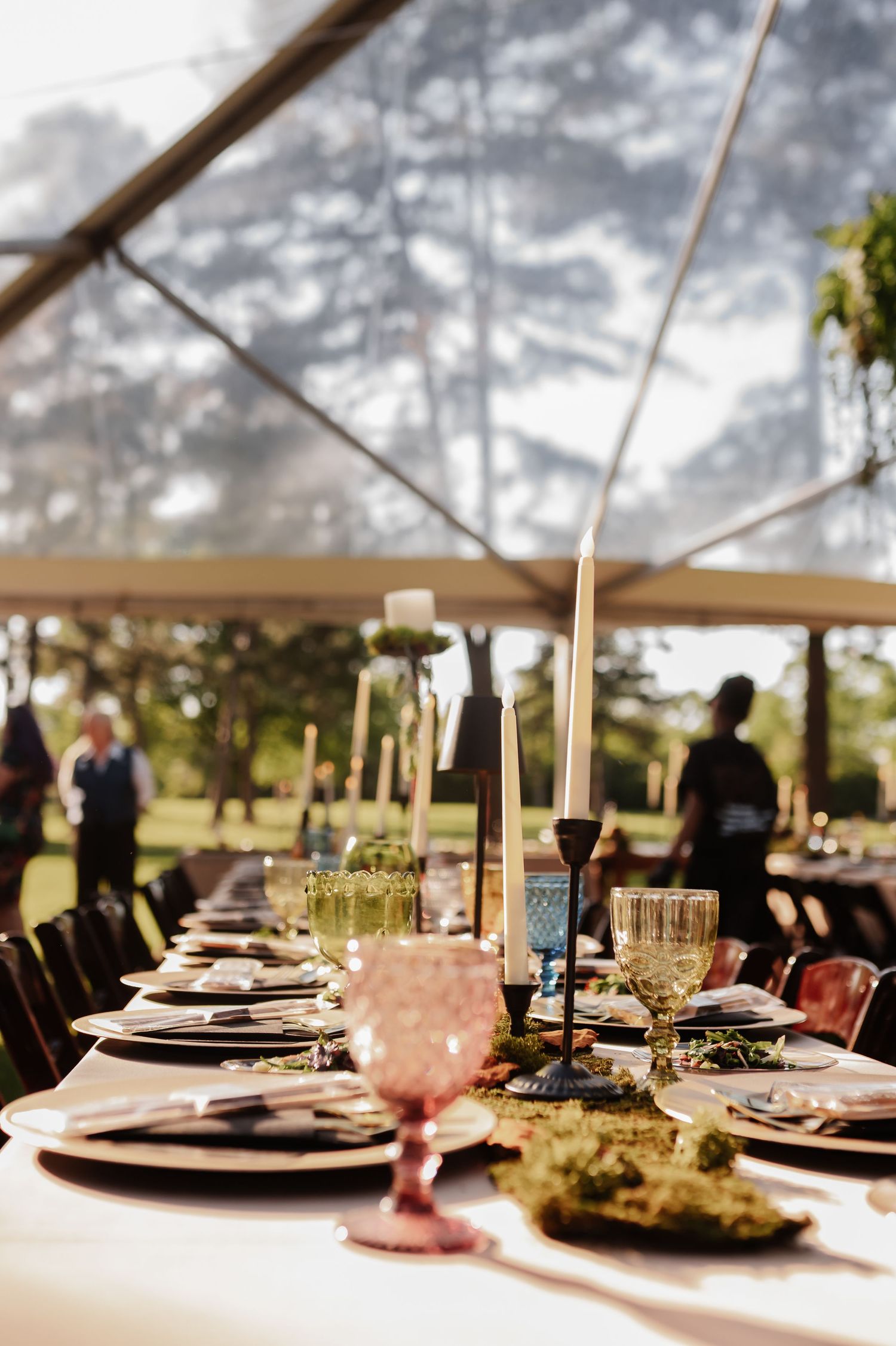 Elegant outdoor wedding reception table set under a clear tent with colorful glassware, candles, and greenery centerpieces.