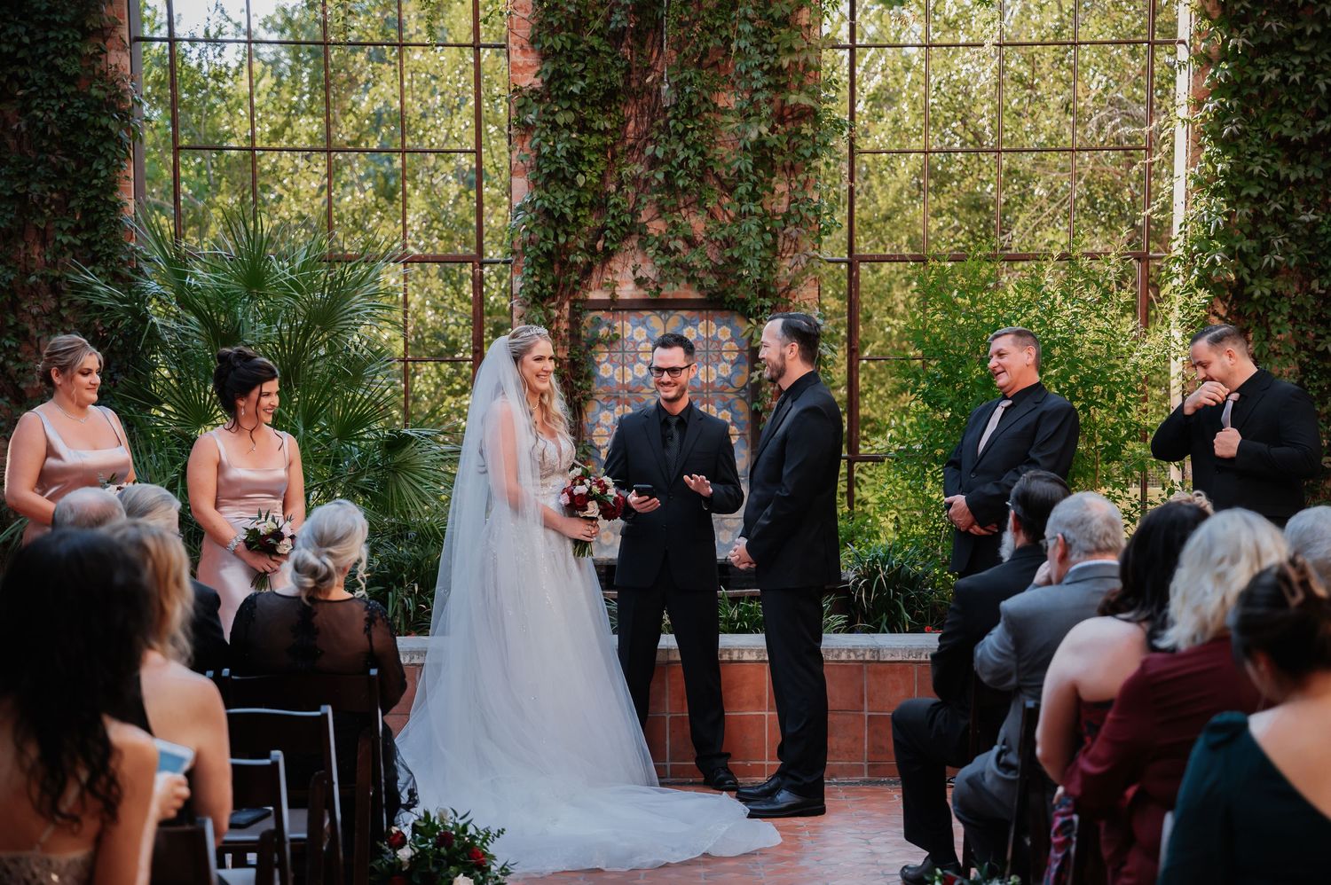 Bride and groom exchange vows at elegant indoor wedding ceremony with large windows and lush greenery backdrop.