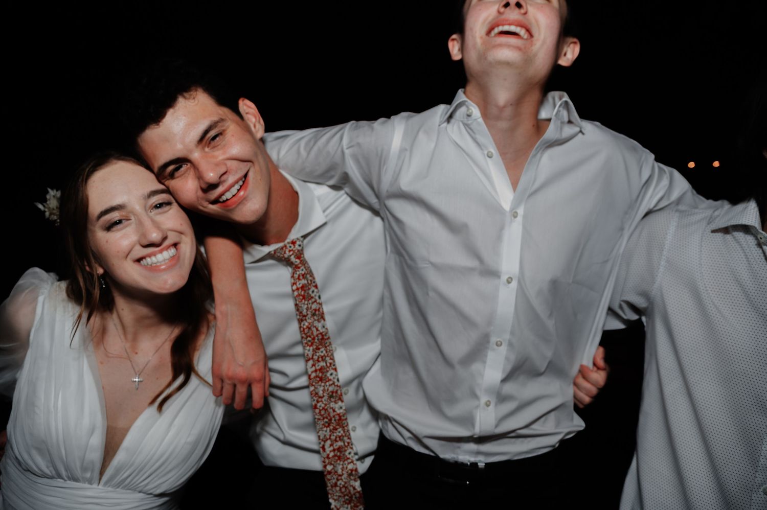 Three young adults smiling and laughing together at a formal evening event, wearing white dress attire.