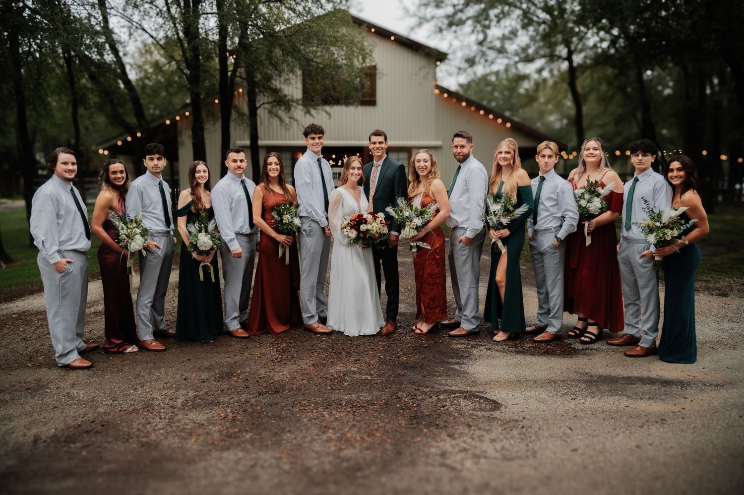 Wedding party poses outdoors in coordinated attire, bridesmaids in burgundy and teal gowns, groomsmen in gray suits.
