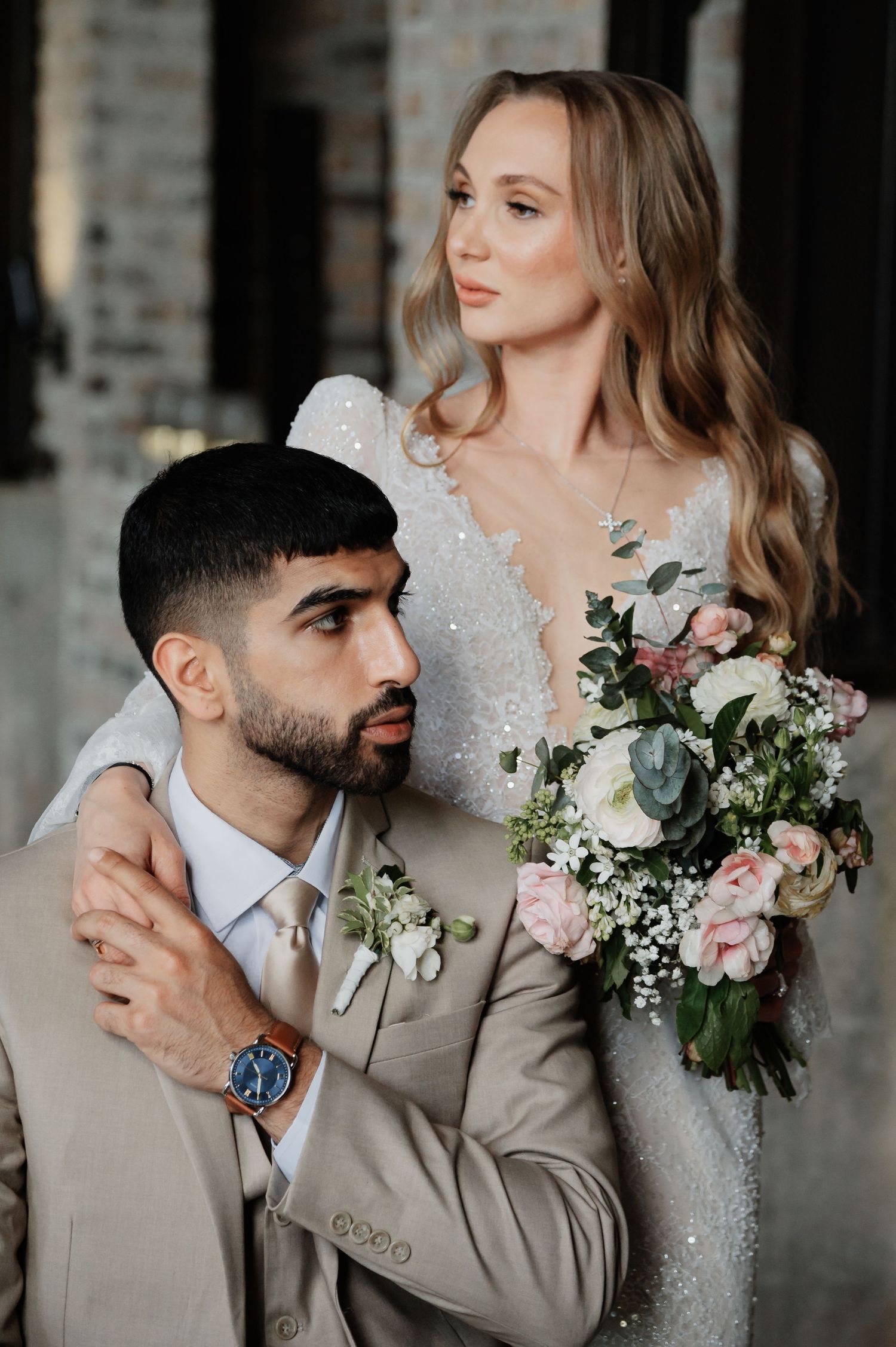 A bride in a sequined gown holds a floral bouquet while embracing a groom in a beige suit with a boutonniere.