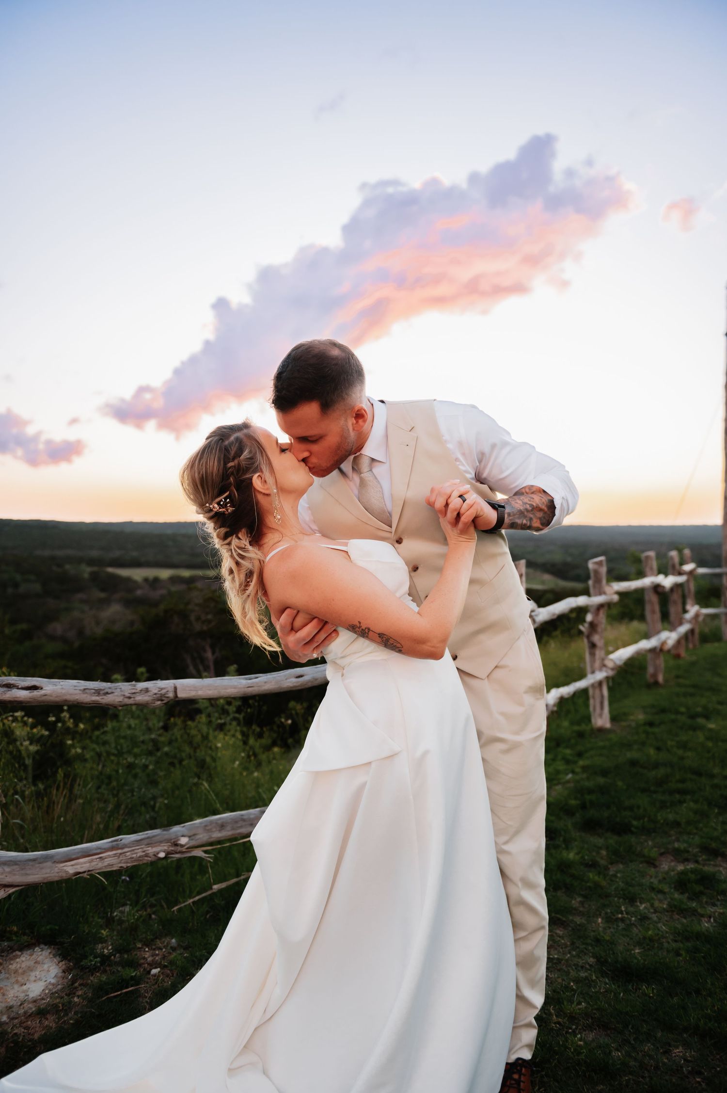 Bride and groom share a romantic sunset kiss in a rustic outdoor wedding setting with a wooden fence backdrop.