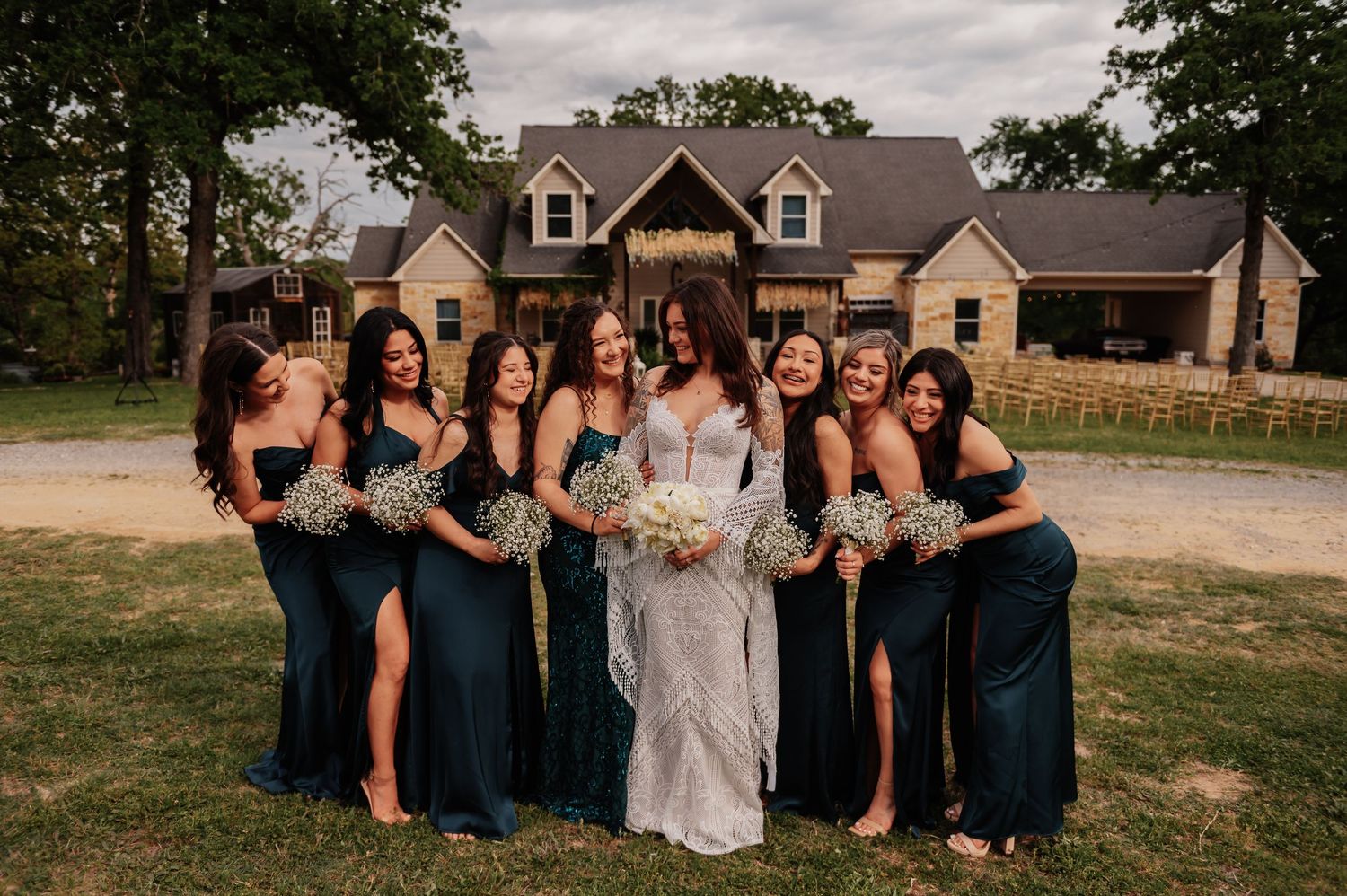 Bride in white lace gown poses with bridesmaids in dark teal dresses holding bouquets outside rustic venue.