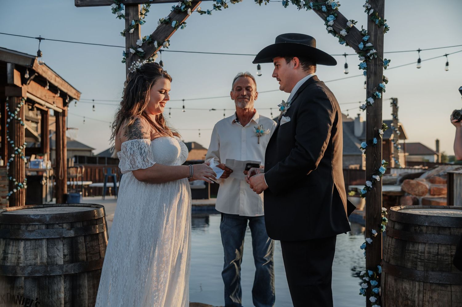 Bride and groom exchange vows outdoors at rustic wedding venue with wooden barrels and string lights at dusk.