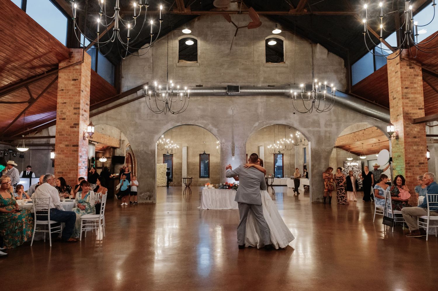 Couple shares first dance in elegant rustic wedding venue with chandeliers, brick columns, and wood floors.