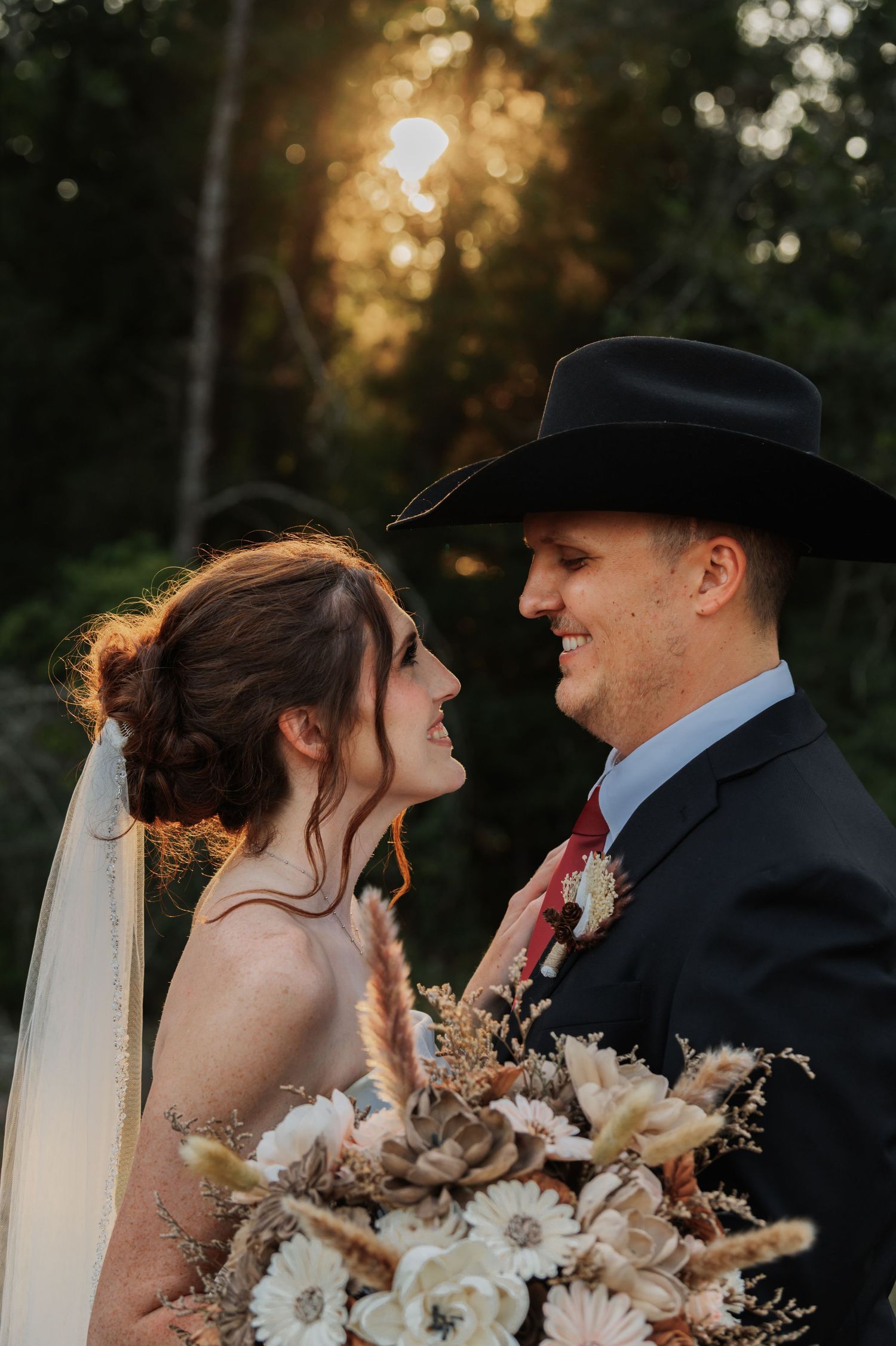 Bride and groom share a romantic golden hour moment, groom wearing a black cowboy hat in the woods.