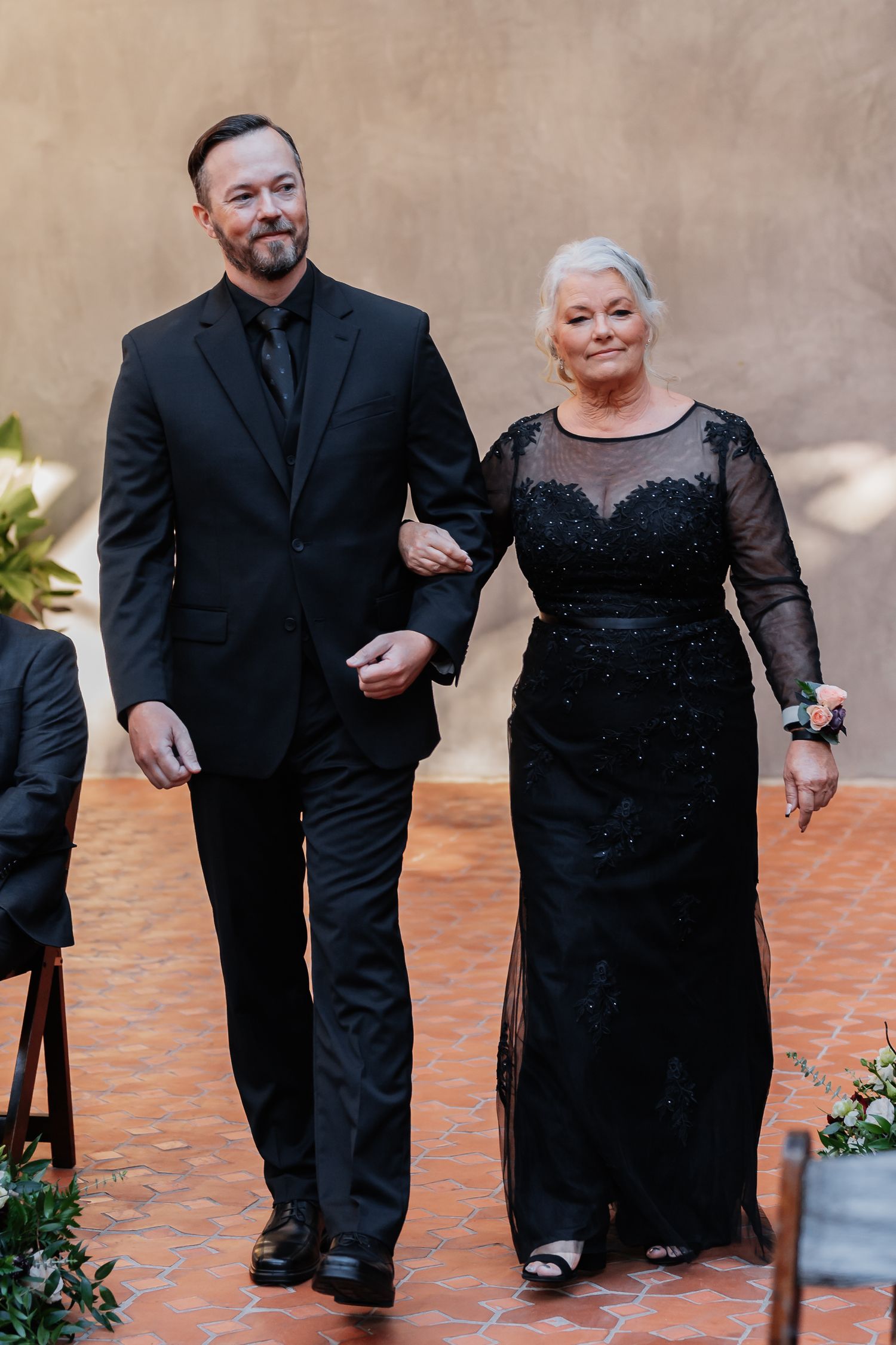 A man in a black suit escorts an older woman in a black lace gown at a formal wedding ceremony.