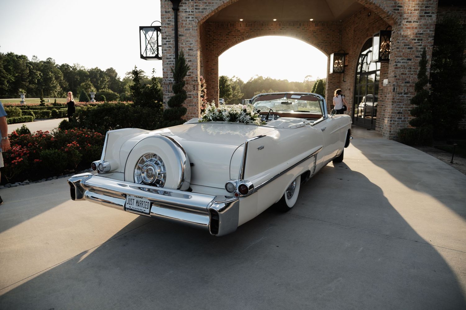Elegant white vintage convertible car decorated with flowers parked outside a brick archway venue.