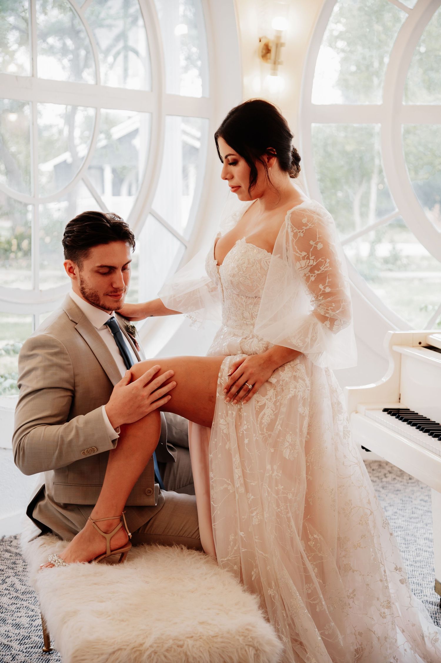 A groom in a gray suit sits while adjusting his bride's garter, as she stands in a lace wedding gown.