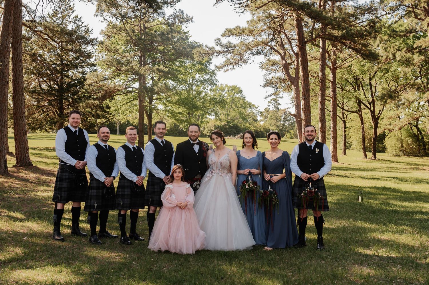 Wedding party in Scottish kilts and blue bridesmaid dresses pose outdoors among tall pine trees with bride and groom.