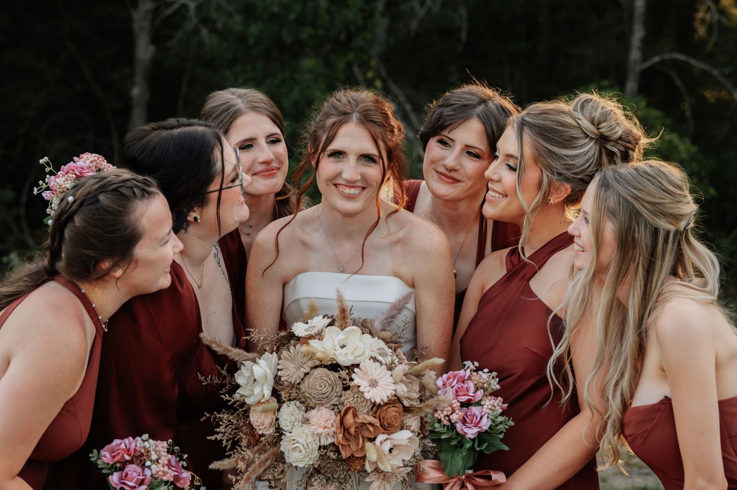 Bride smiles surrounded by bridesmaids in burgundy dresses holding elegant floral bouquets outdoors.