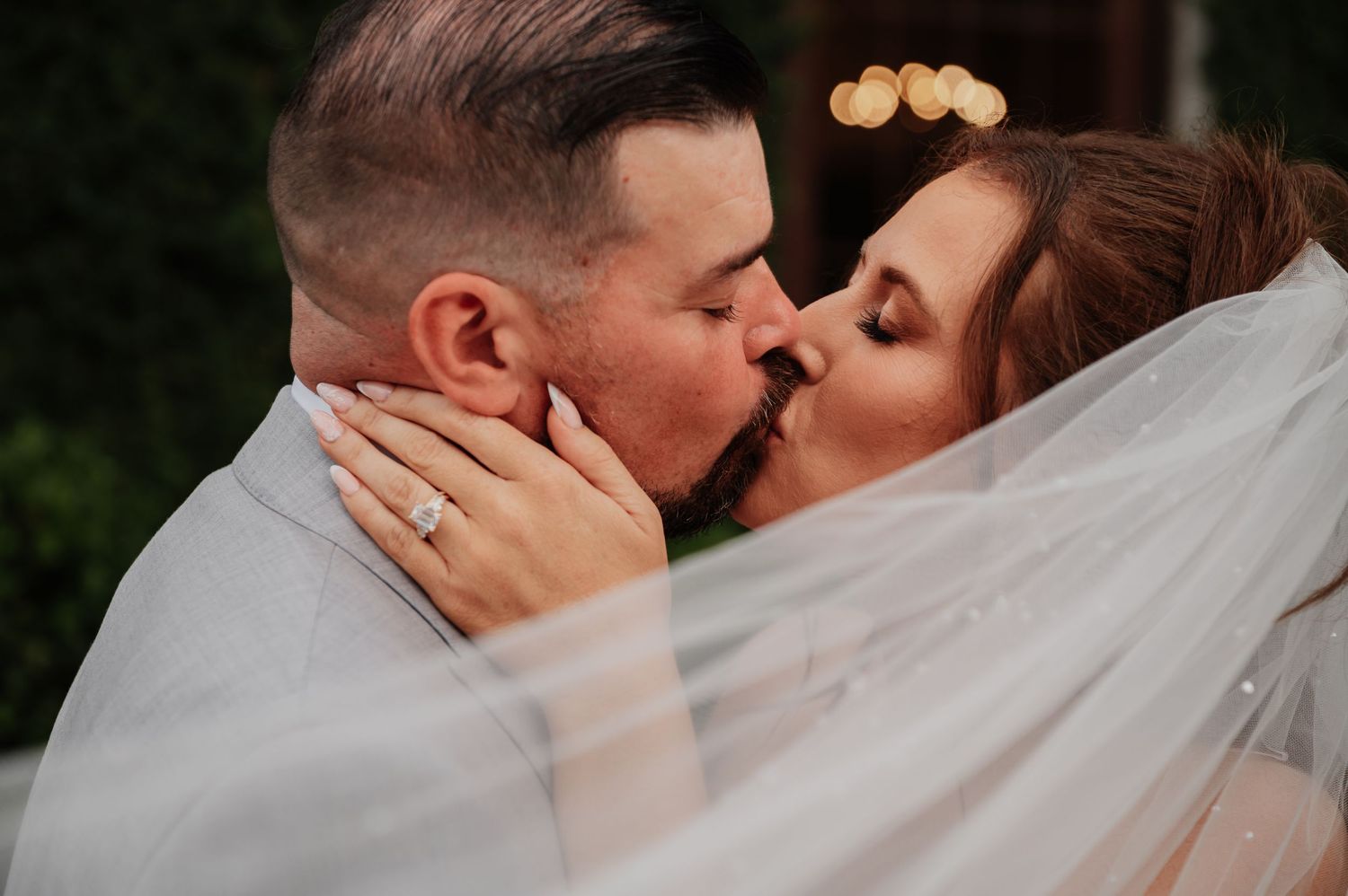 A bride and groom share a romantic kiss under a flowing white veil during their wedding ceremony outdoors.
