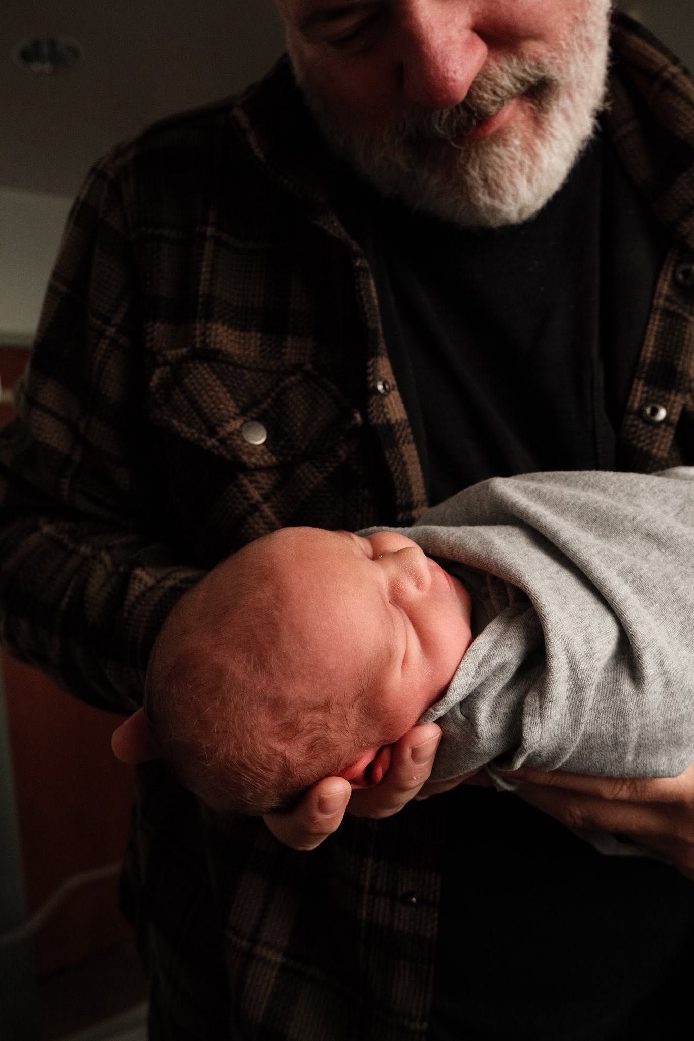 Bearded grandfather tenderly cradling a sleeping newborn baby wrapped in a gray blanket.