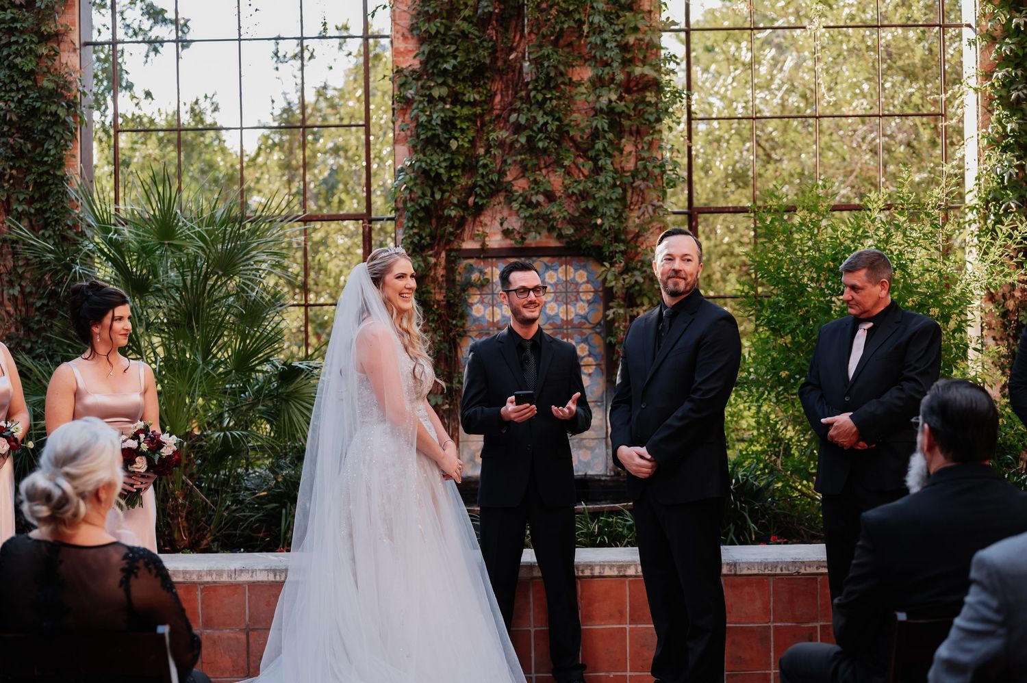 Bride in white gown and veil stands with groom and officiant during outdoor wedding ceremony at rustic brick venue.