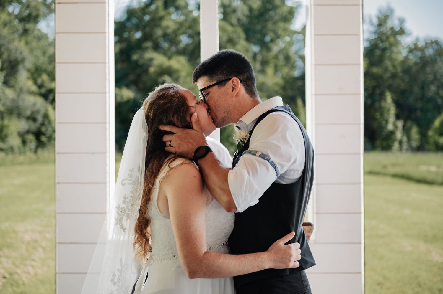 Bride and groom share a romantic kiss between white pillars outdoors on their wedding day.