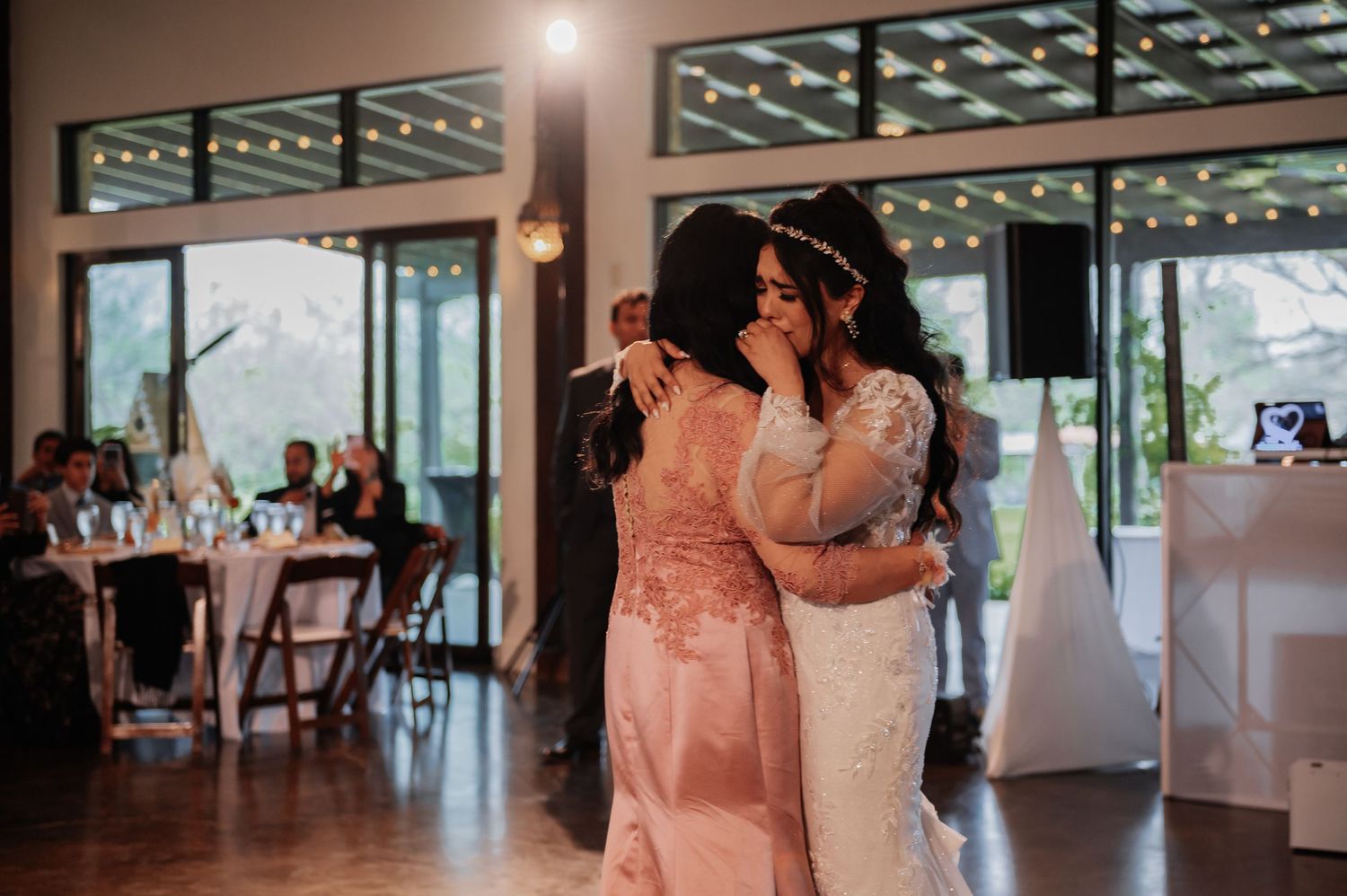 Two women share an emotional embrace on the dance floor during a wedding reception with string lights.