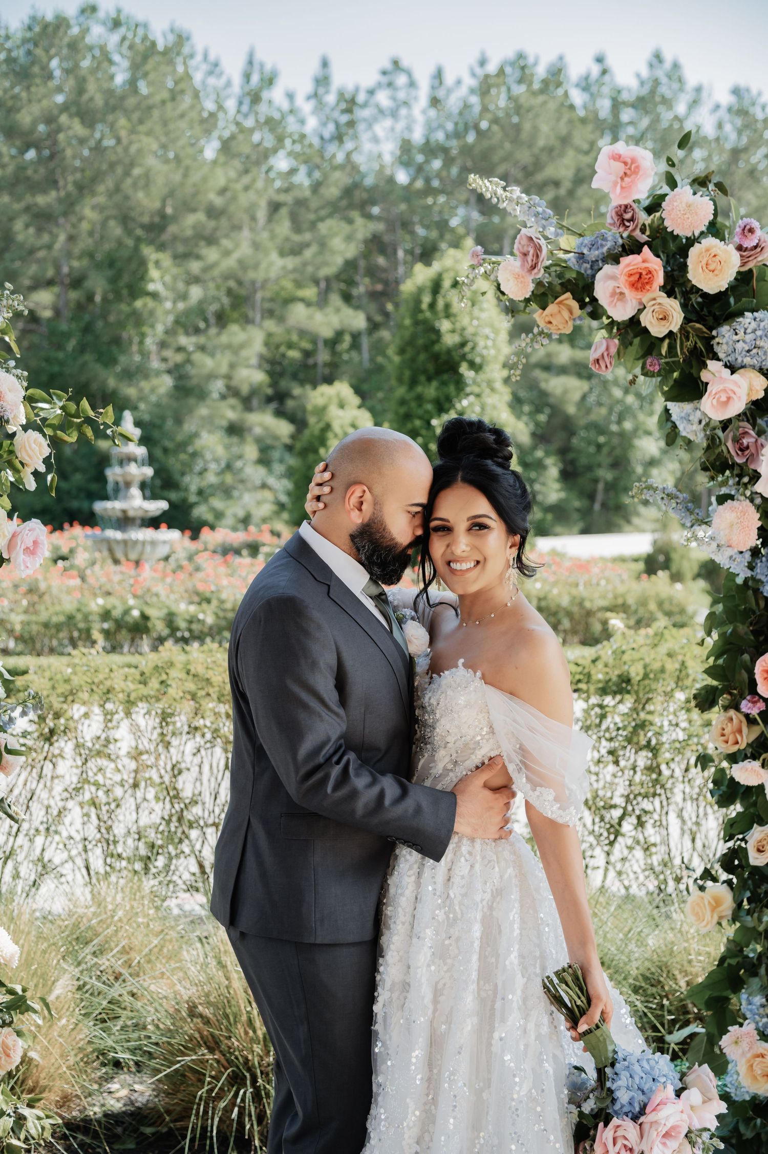 Bride and groom embrace under a floral arch with pink and white blooms at an outdoor garden wedding ceremony.
