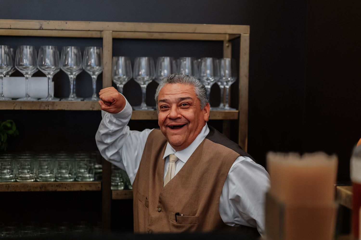 Cheerful waiter in vest raises fist triumphantly behind bar with wine glasses on wooden shelves.