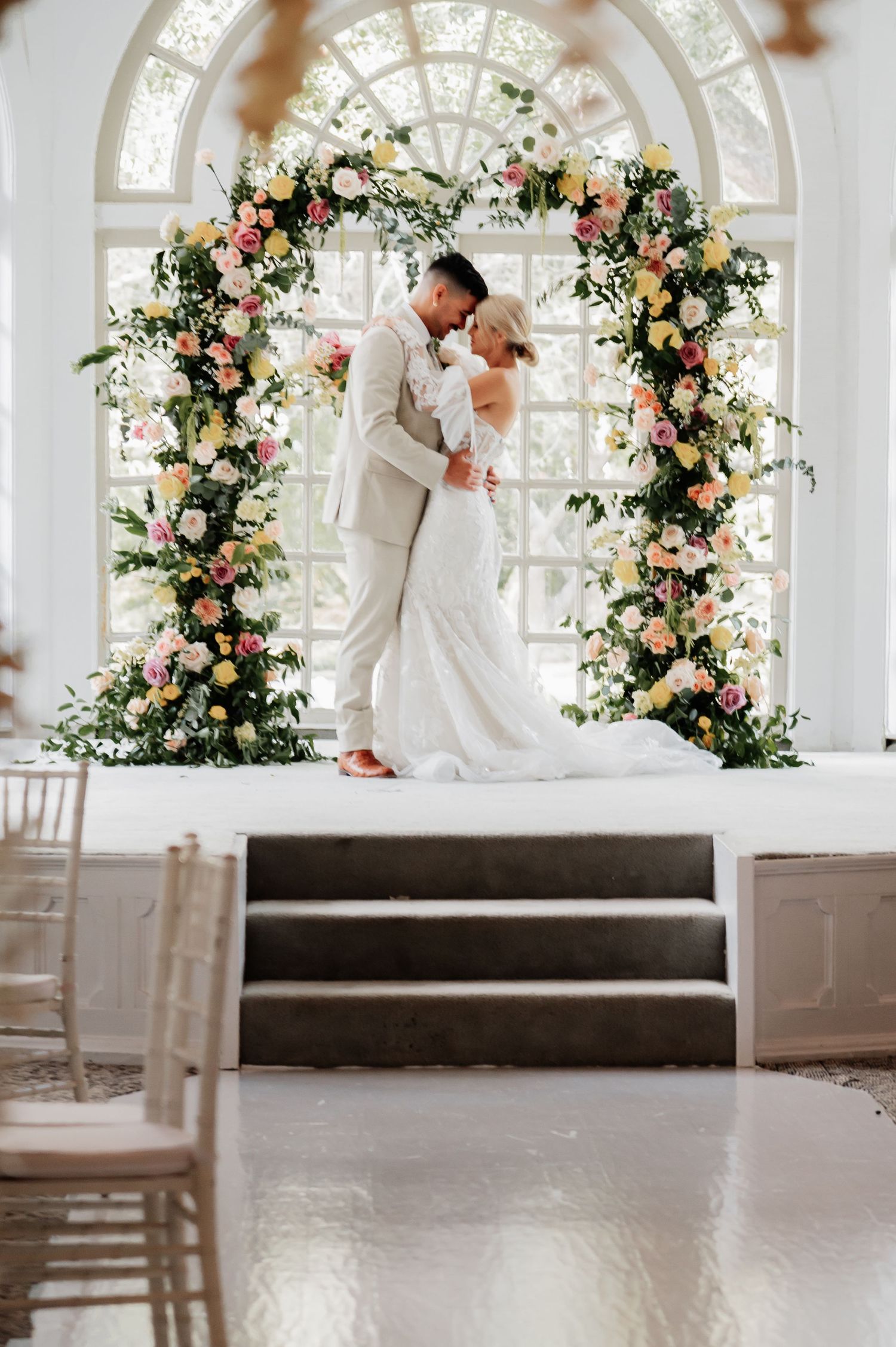 Bride and groom embrace under a floral arch with pink, yellow, and white blooms in an elegant indoor wedding venue.