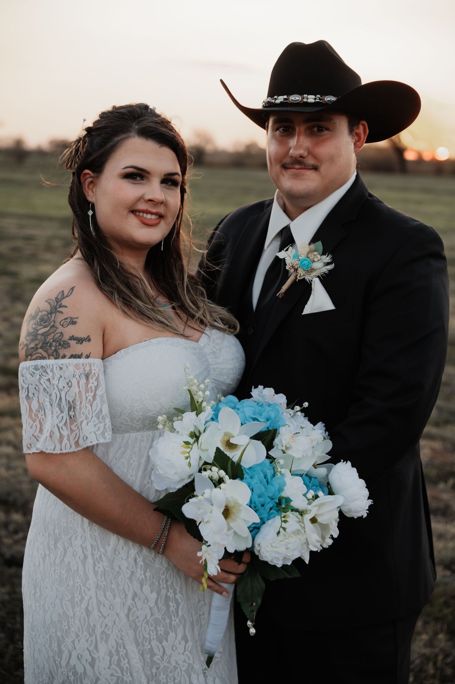 Bride in white lace dress holding blue and white bouquet stands with groom in black suit and cowboy hat at sunset.