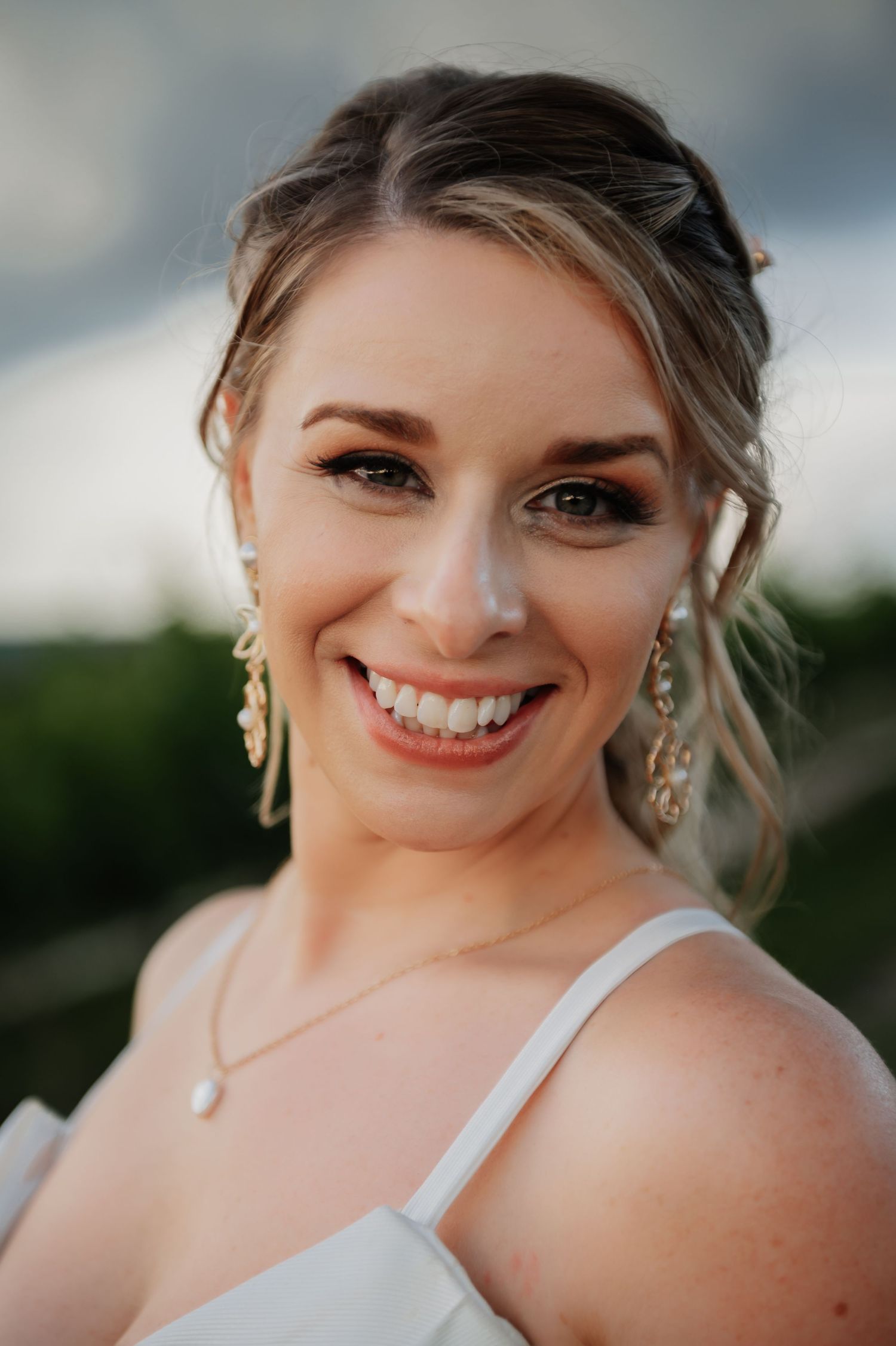 Smiling bride with updo hairstyle, gold drop earrings, and pearl necklace wearing a white dress outdoors.