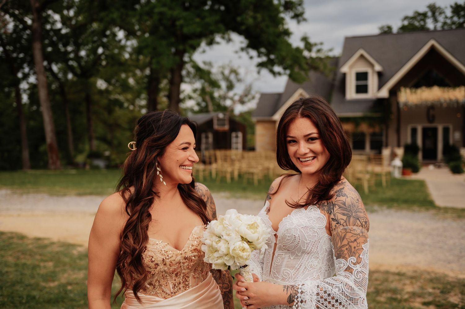 Two women smile outdoors at a wedding, one in a lace gown holding white flowers, near a rustic venue.