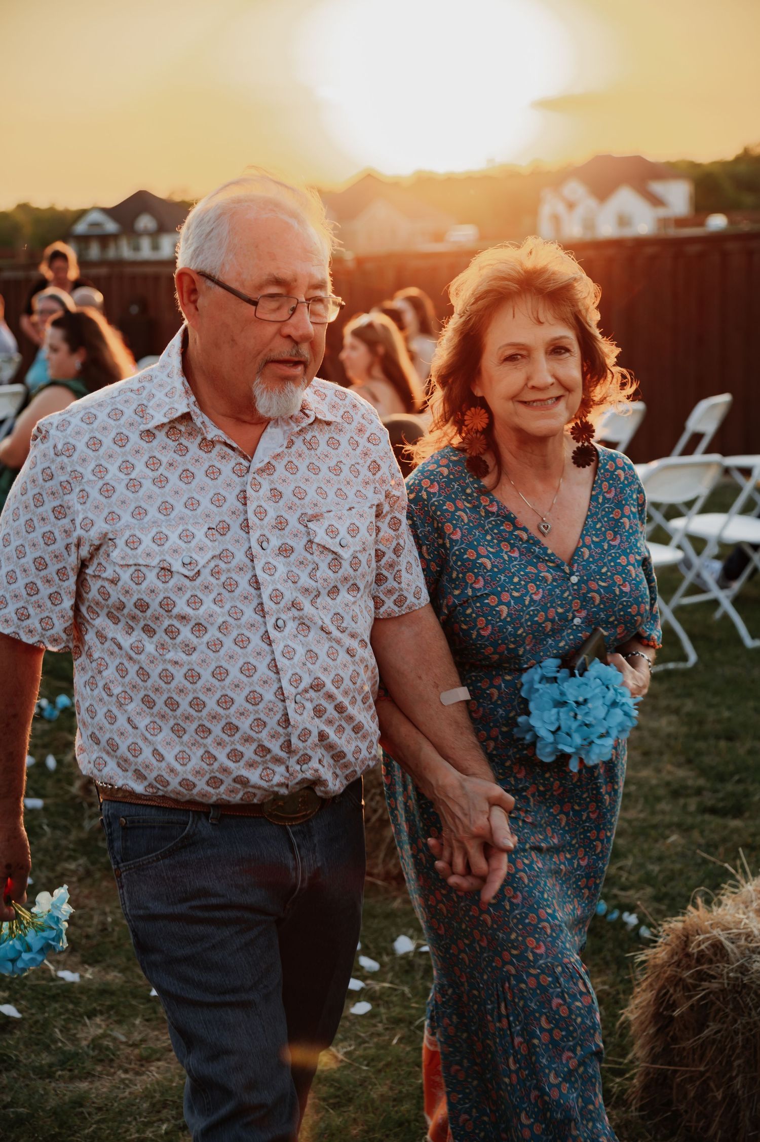 Elegant older couple walking hand-in-hand at outdoor backyard wedding ceremony during golden hour sunset.
