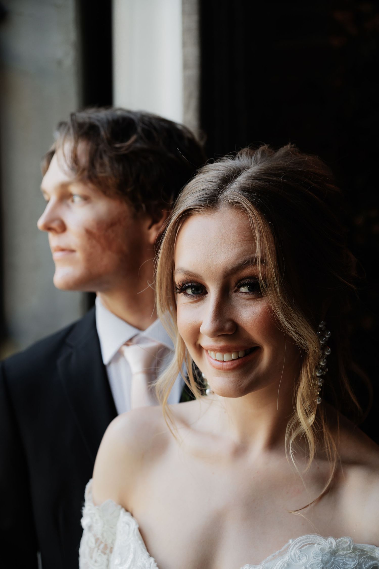 Smiling bride in off-shoulder lace gown with elegant earrings, groom in dark suit with pink tie softly blurred behind her.