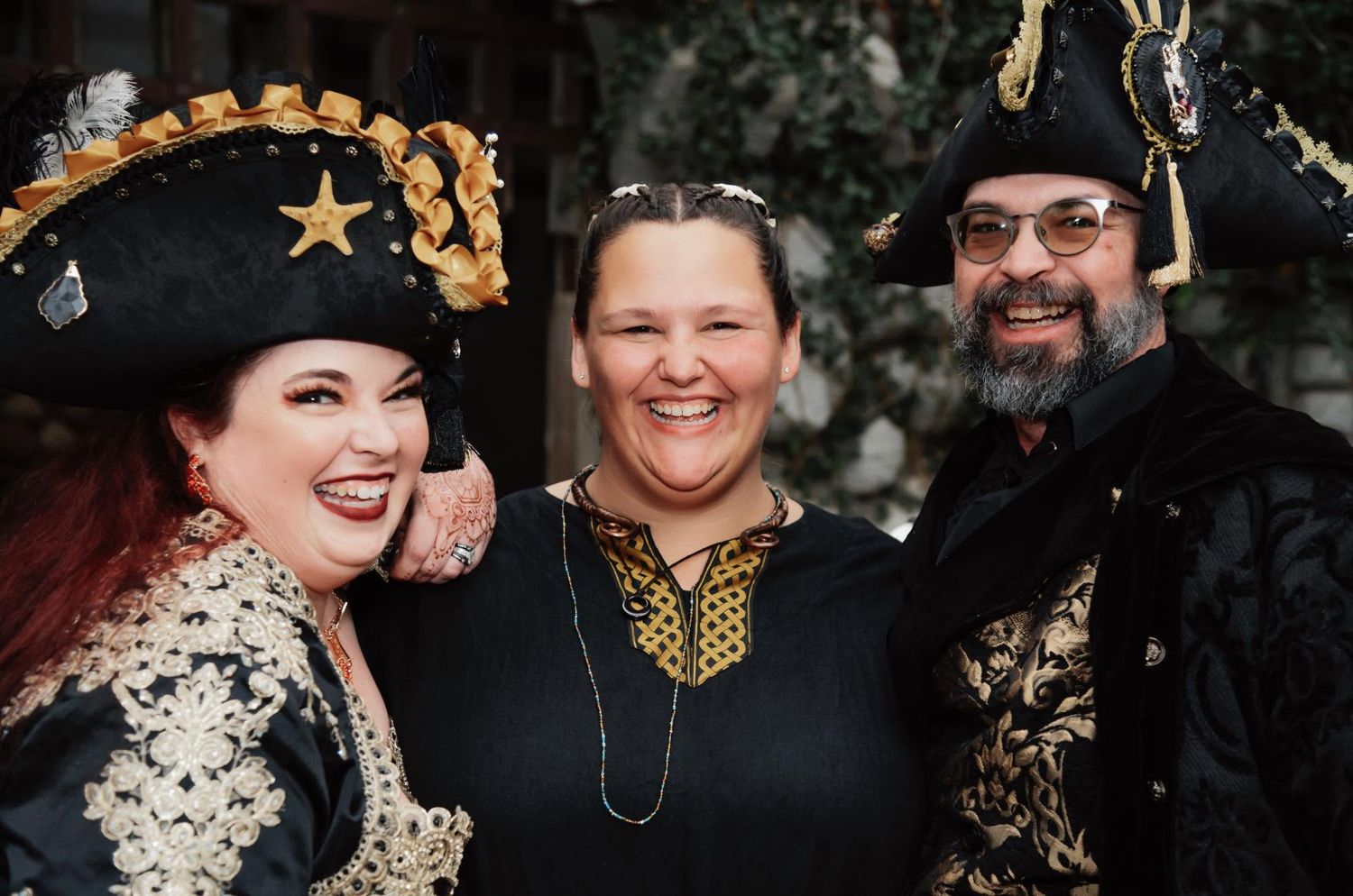 Three people in pirate costumes smile together, two wearing ornate black and gold hats at a costume event.