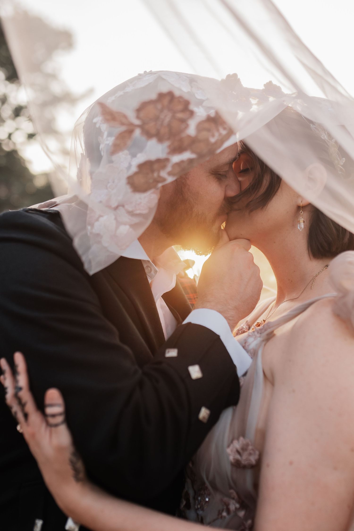 Bride and groom share a romantic kiss under a floral lace veil with golden sunset light glowing between them.
