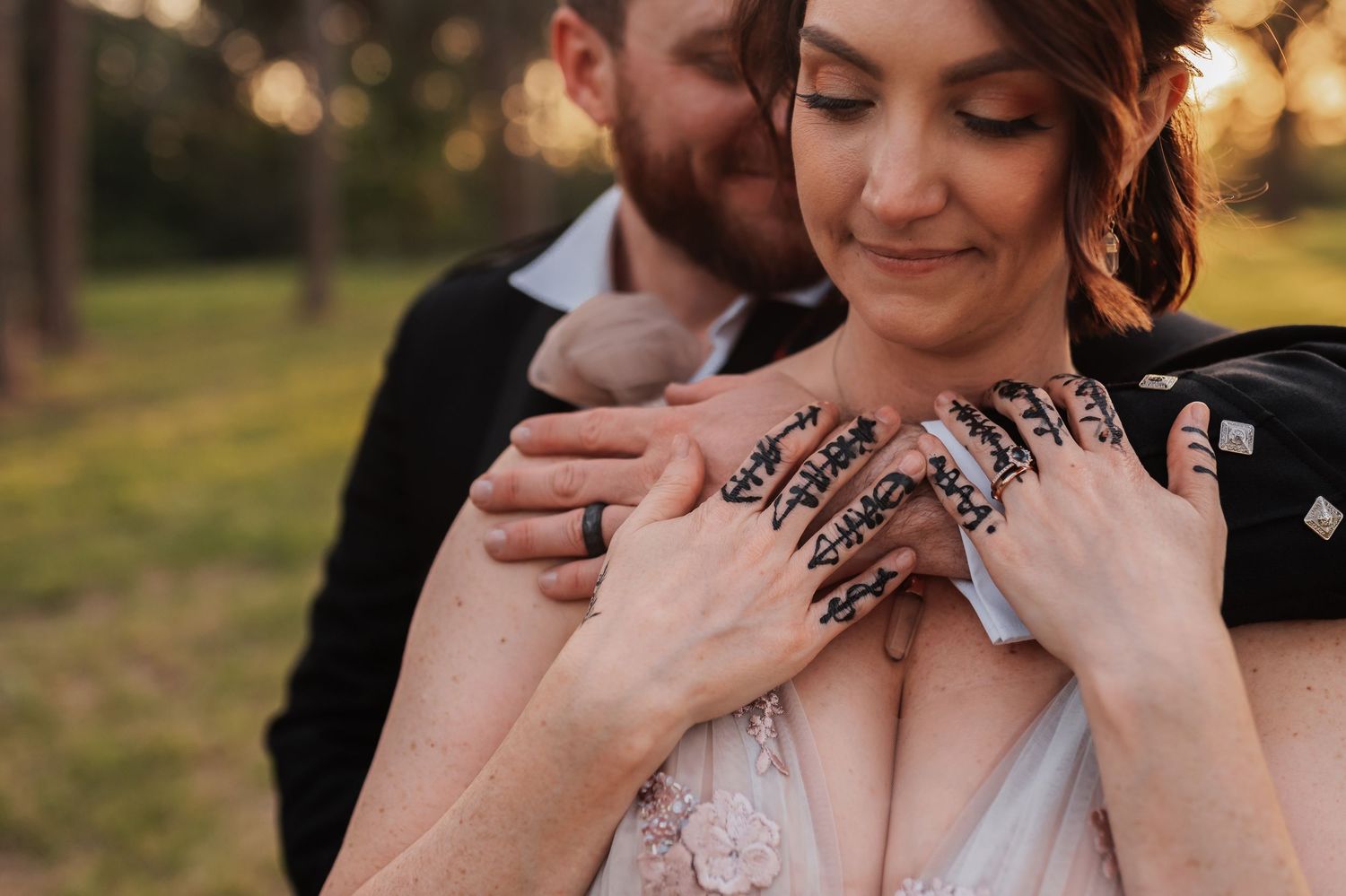 Bride with henna hands embraced by groom in black suit during golden hour outdoor wedding photo.