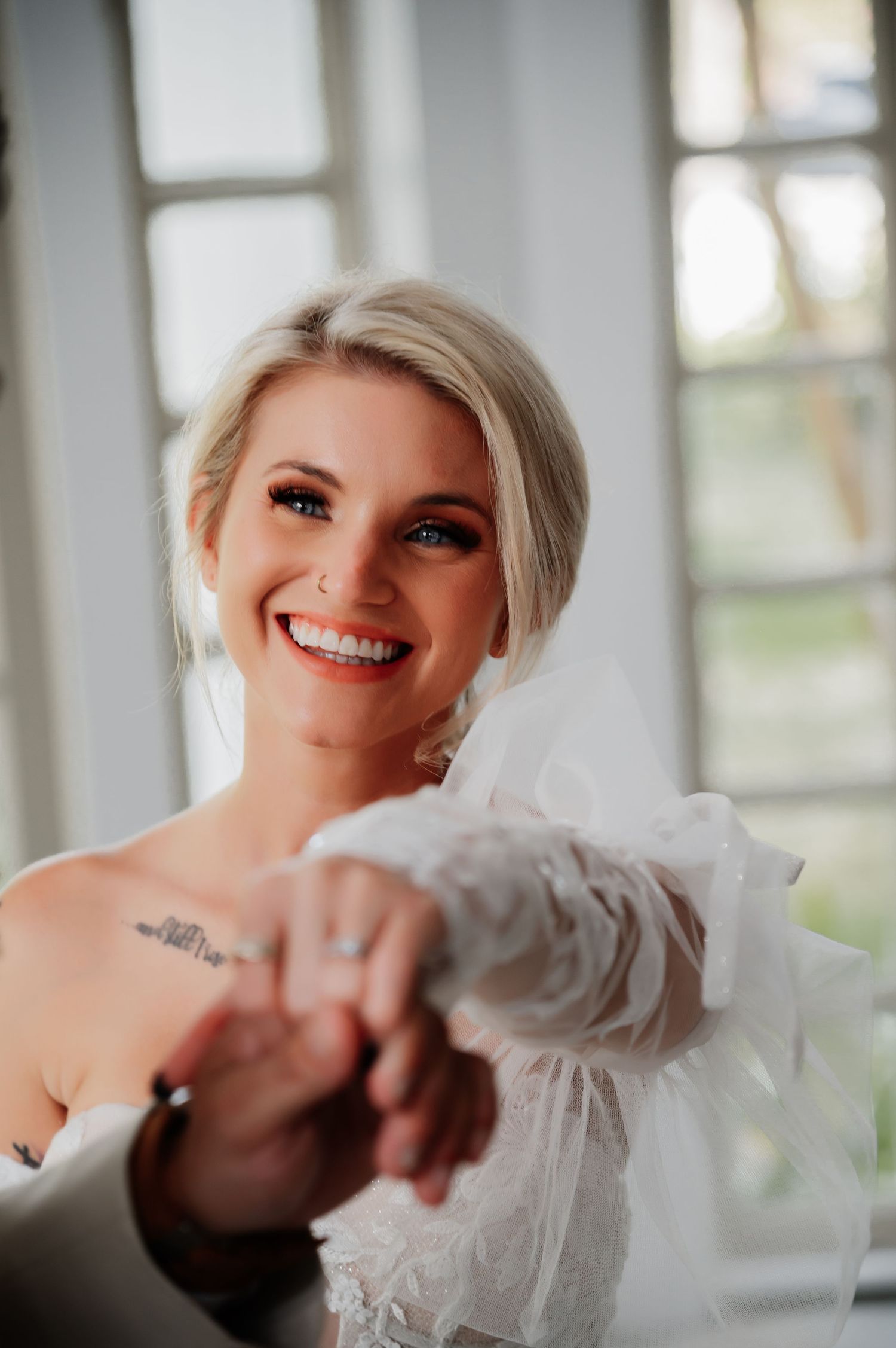 Smiling blonde bride in white lace gown holds hands with someone, wearing a veil and tattoo visible on her chest.