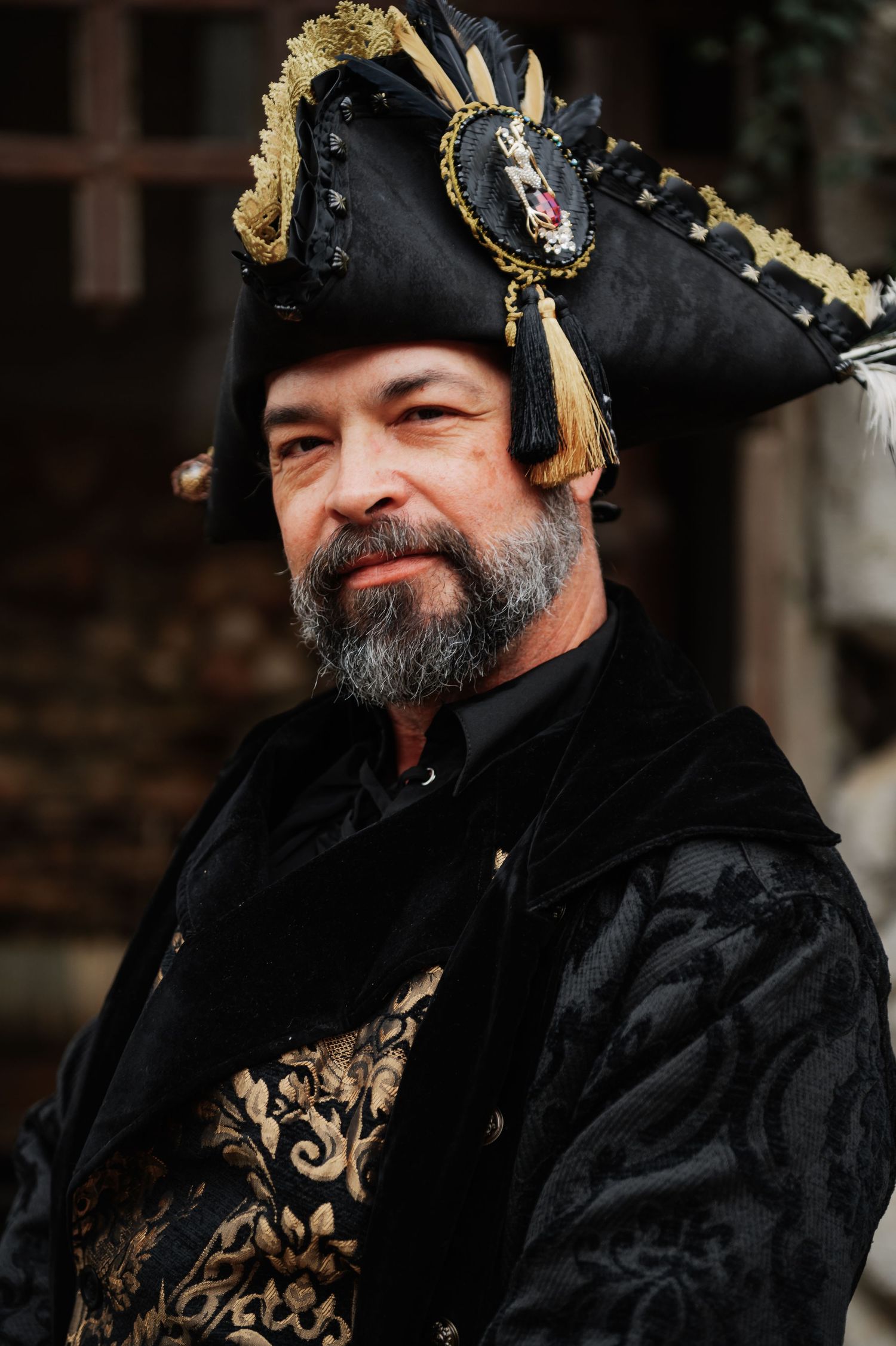 Man in ornate black and gold pirate costume with tricorn hat and beard posing outdoors.