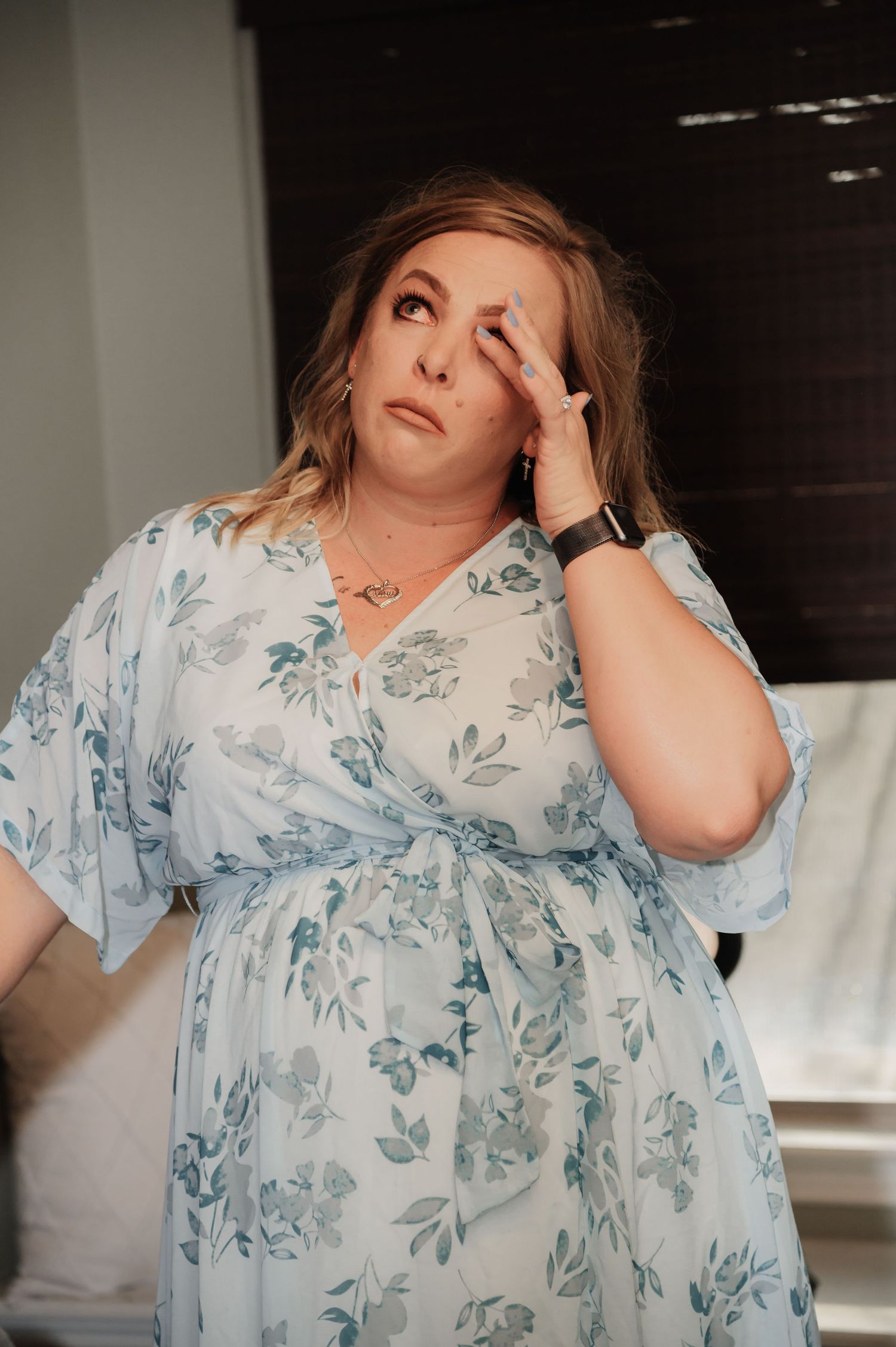 Woman in light blue floral dress with hand raised to face, looking upward thoughtfully indoors.