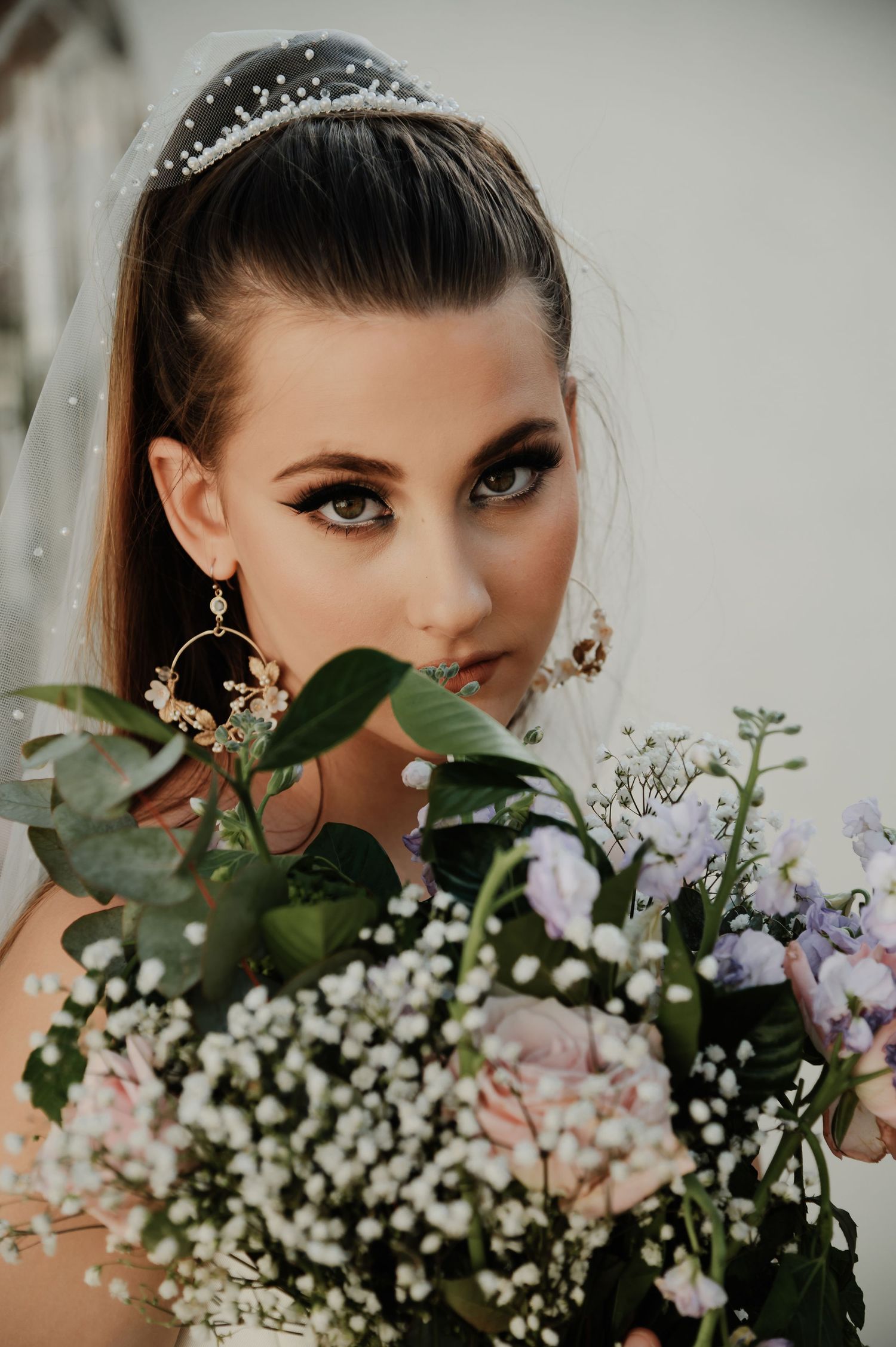 Close-up of bride with dramatic eye makeup, tiara, veil, and gold hoop earrings holding pastel floral bouquet.