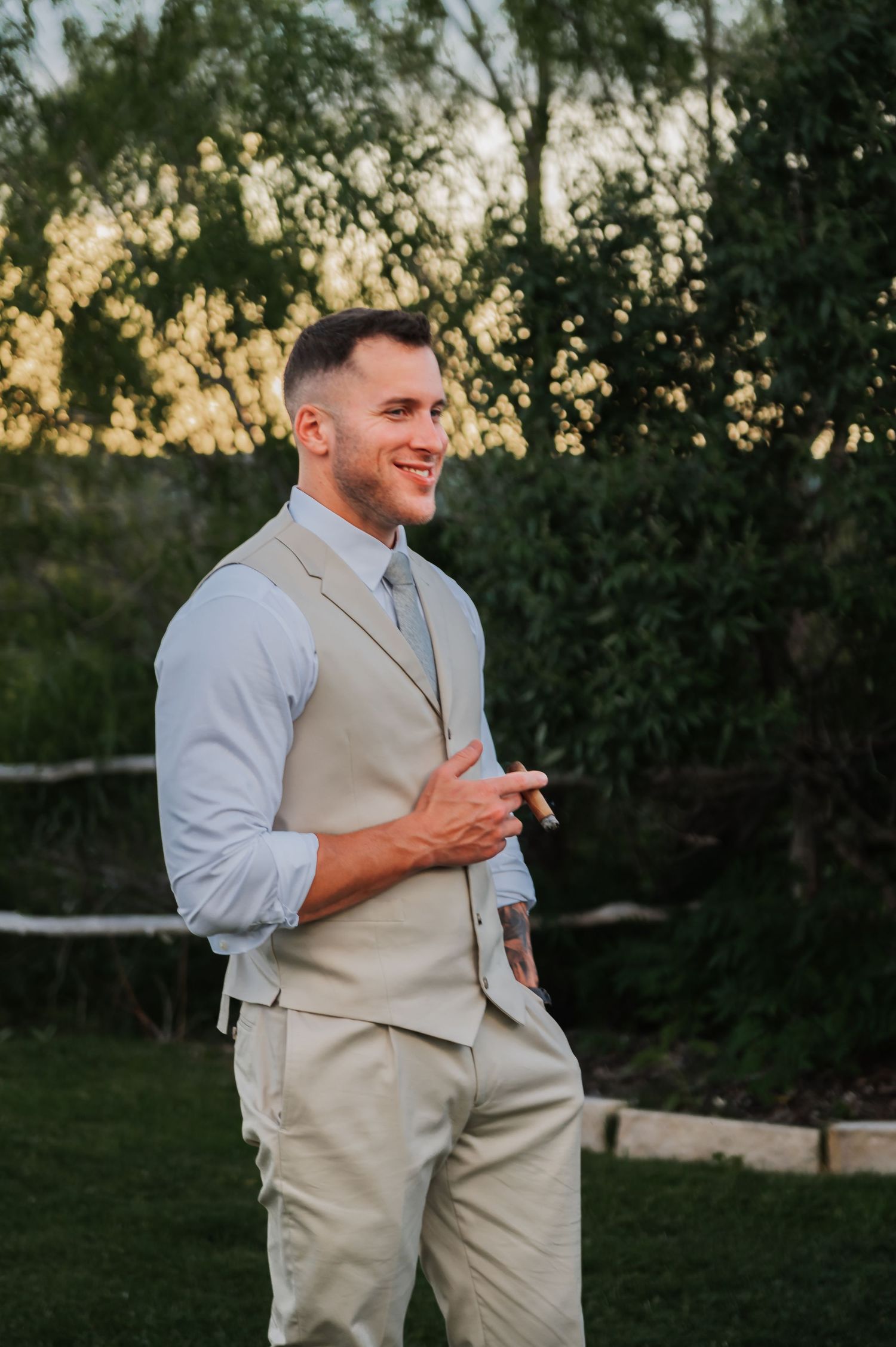 Smiling groom in tan vest and light blue shirt stands outdoors at golden hour with lush green trees behind him.