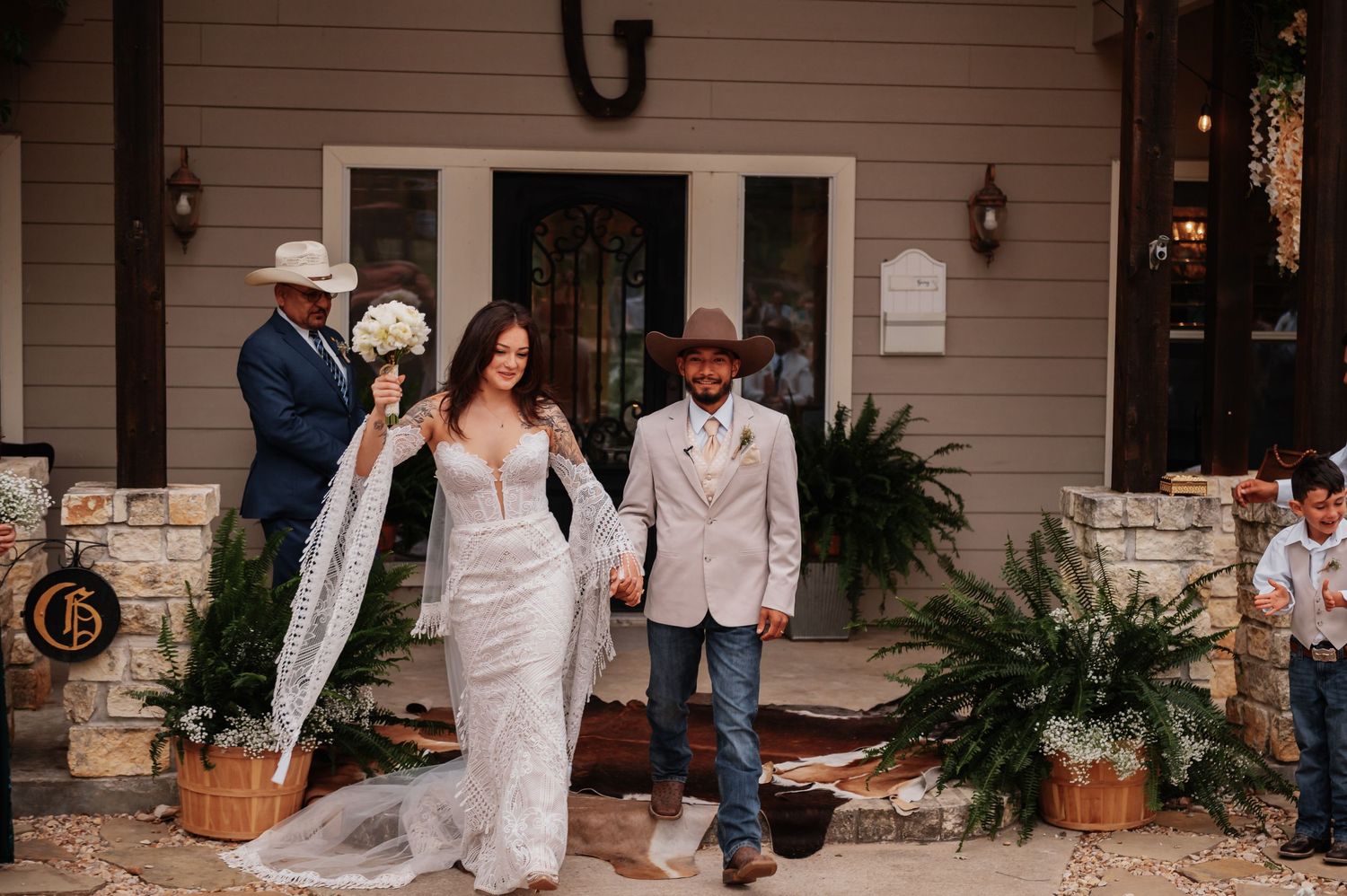 Bride in lace gown walks with groom in tan blazer and cowboy hat outside rustic wedding venue.