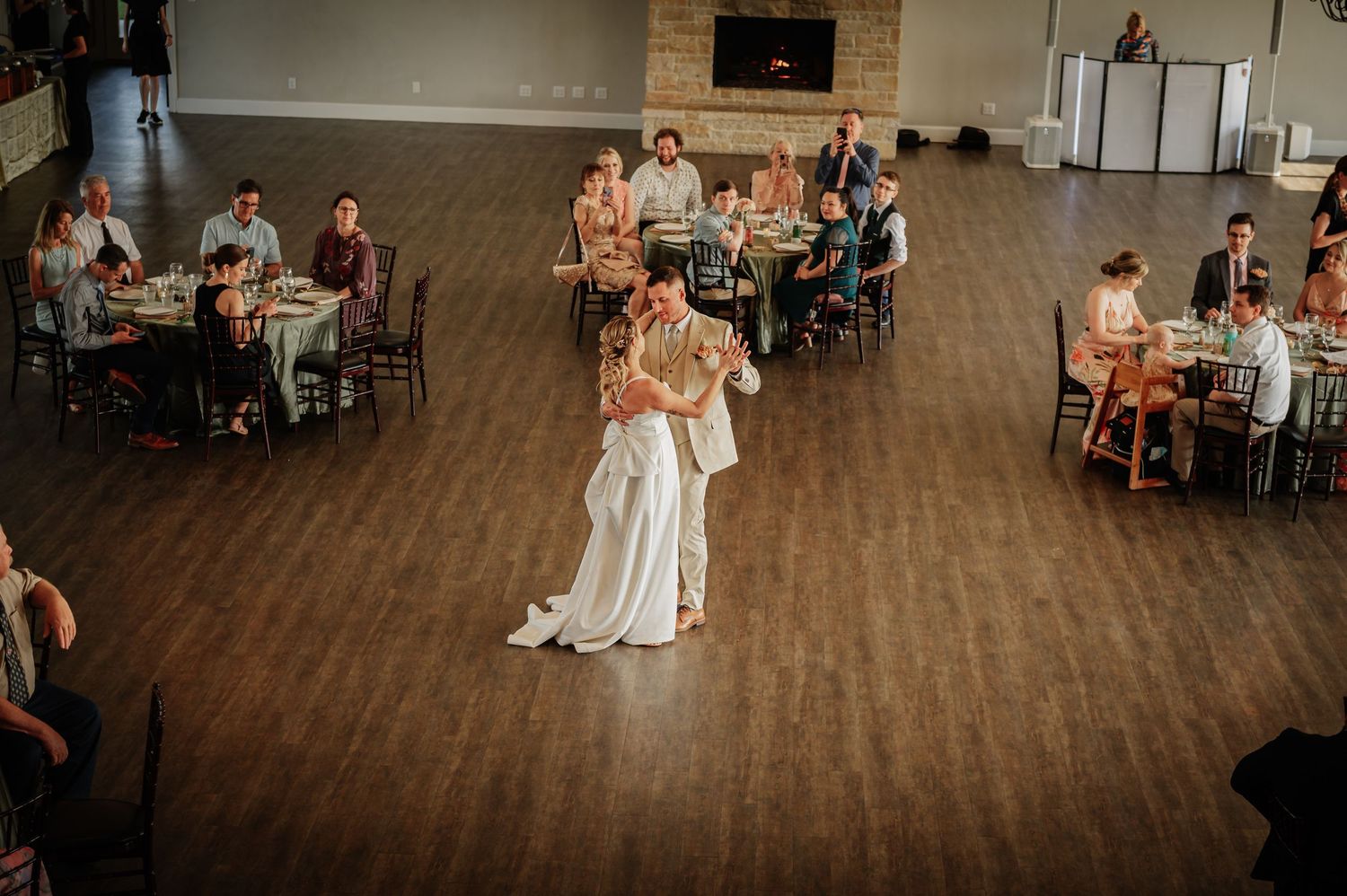 Bride and groom share their first dance on a wooden floor surrounded by seated wedding guests in an elegant reception hall.