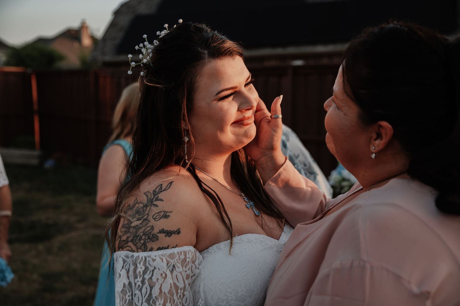 Emotional bride in white lace dress with floral crown being comforted at outdoor wedding reception at sunset.