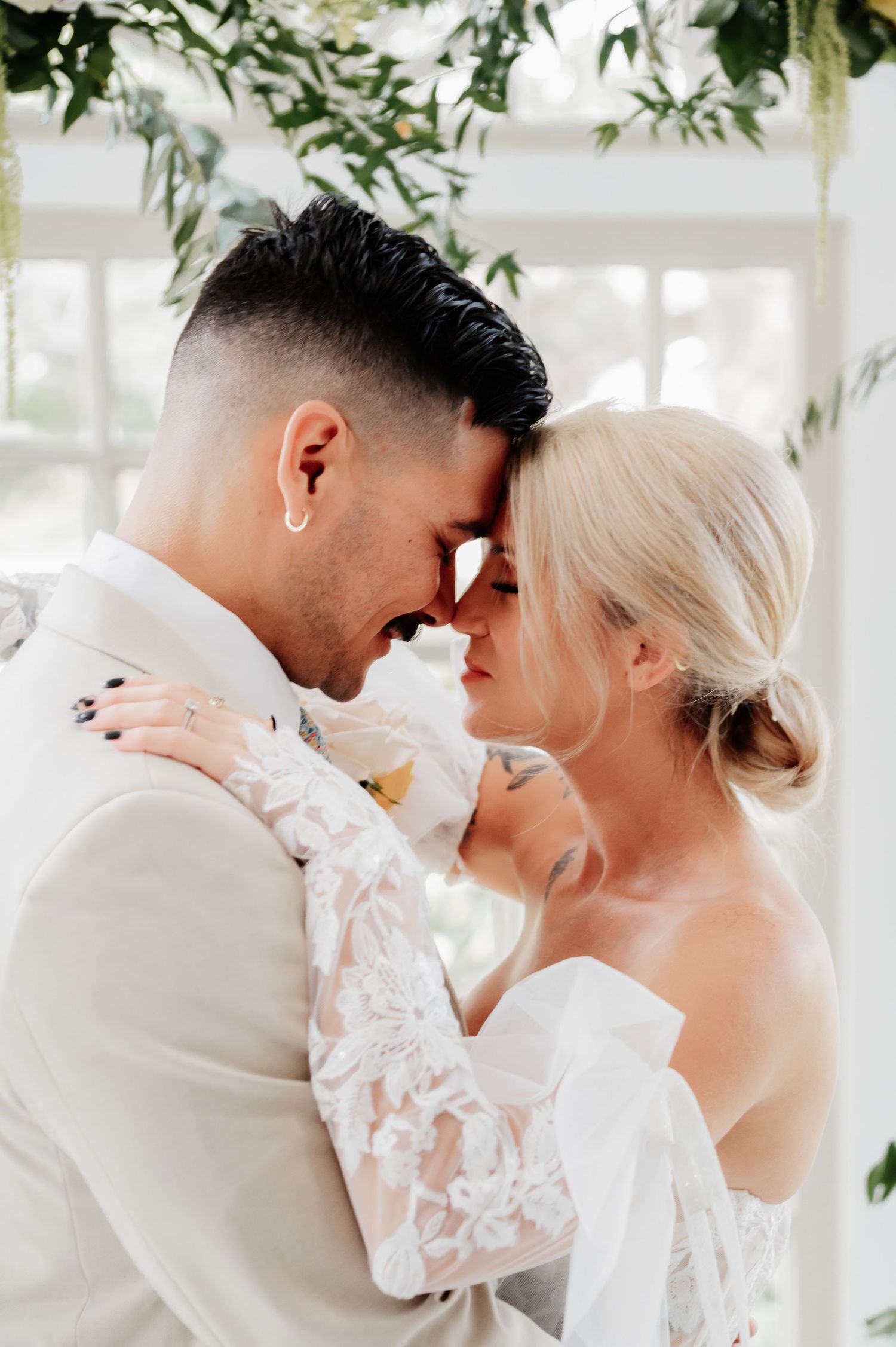 A bride and groom in wedding attire touch foreheads tenderly under a floral arch in a bright, elegant venue.